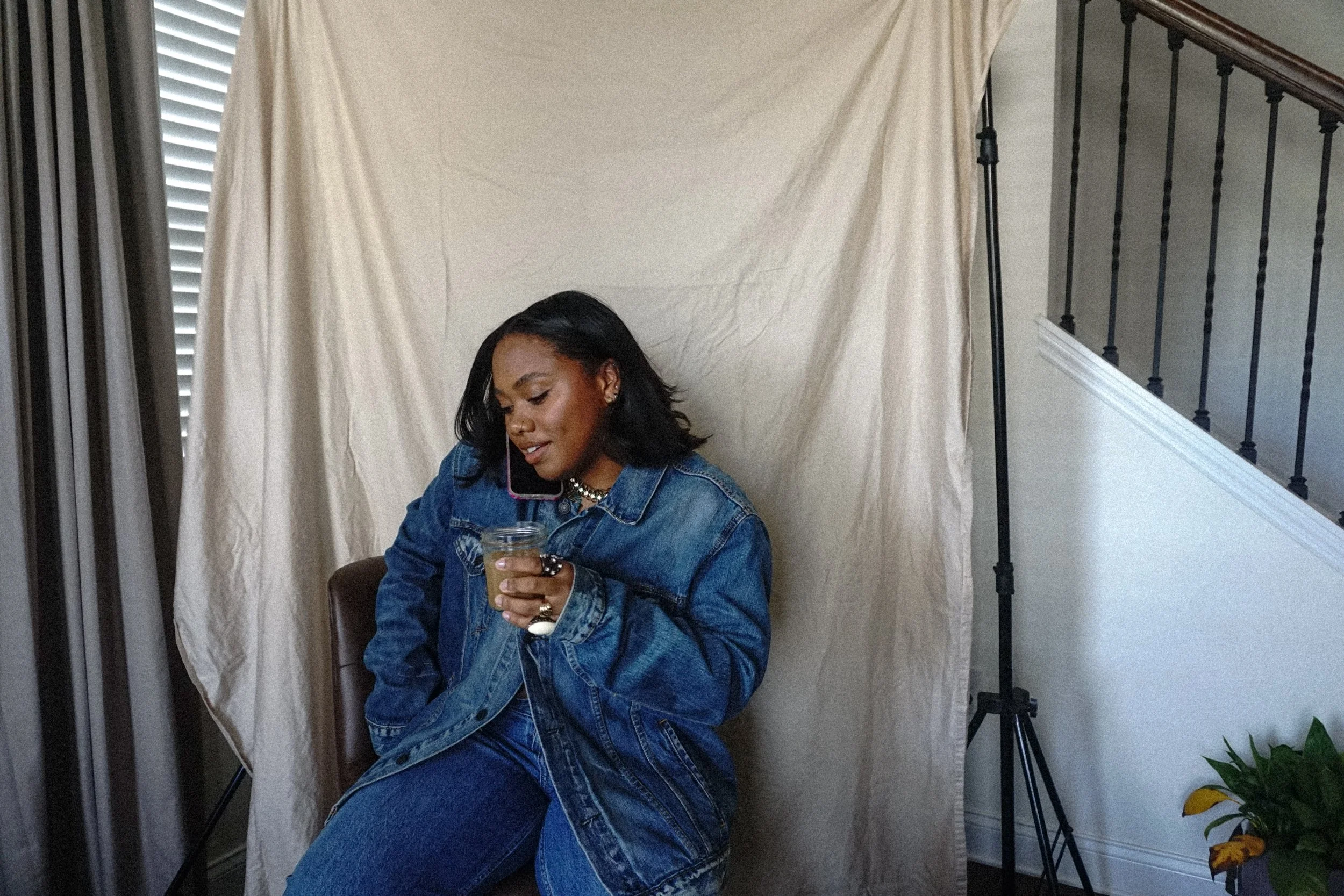 A woman sitting on a chair, holding a glass jar, talking on her cell phone near a beige backdrop and window blinds, with a plant in the corner.