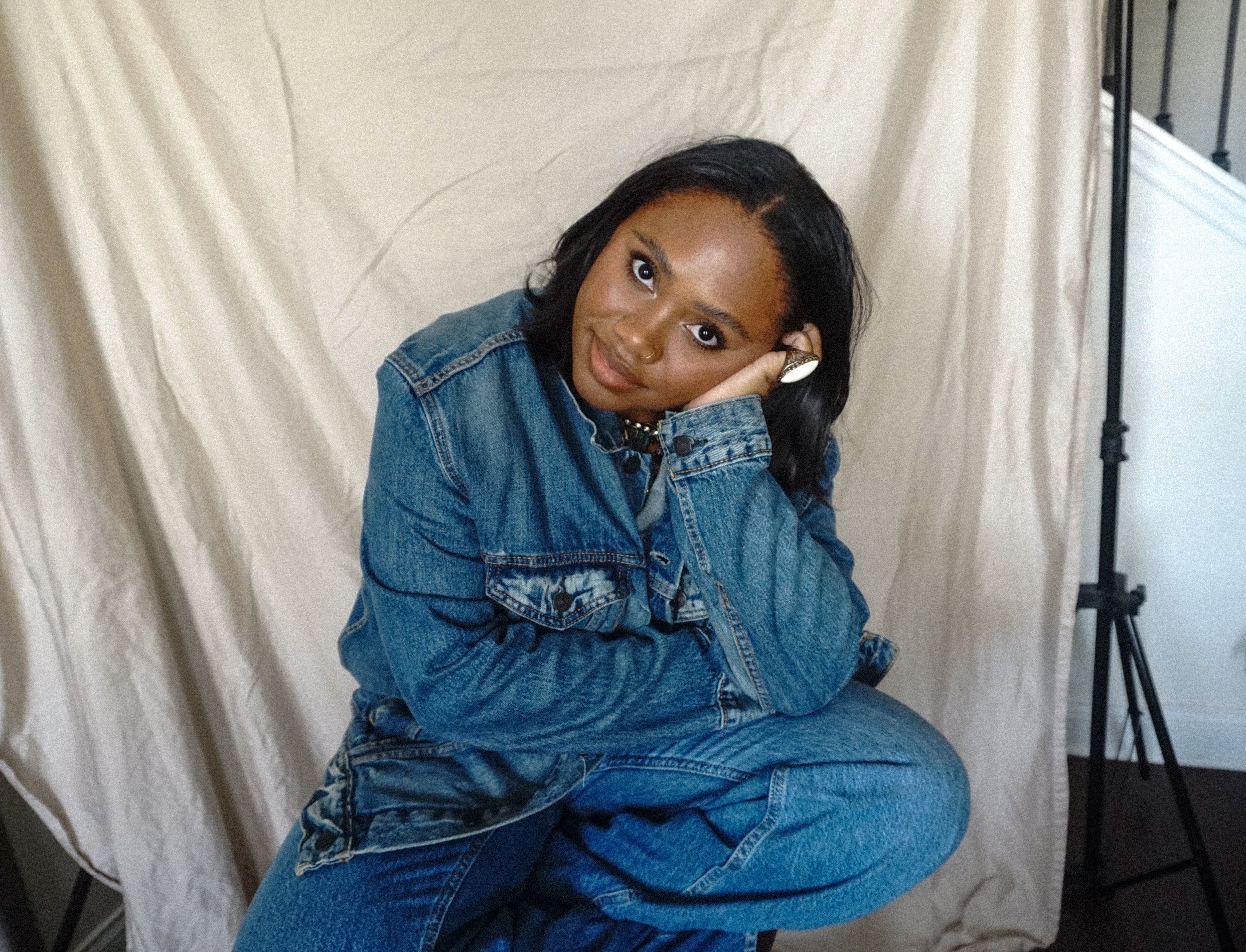 A woman with dark hair and medium brown skin wearing a denim jacket and pants, sitting with her chin resting on her right hand, looking at the camera against a light fabric backdrop and light stand.