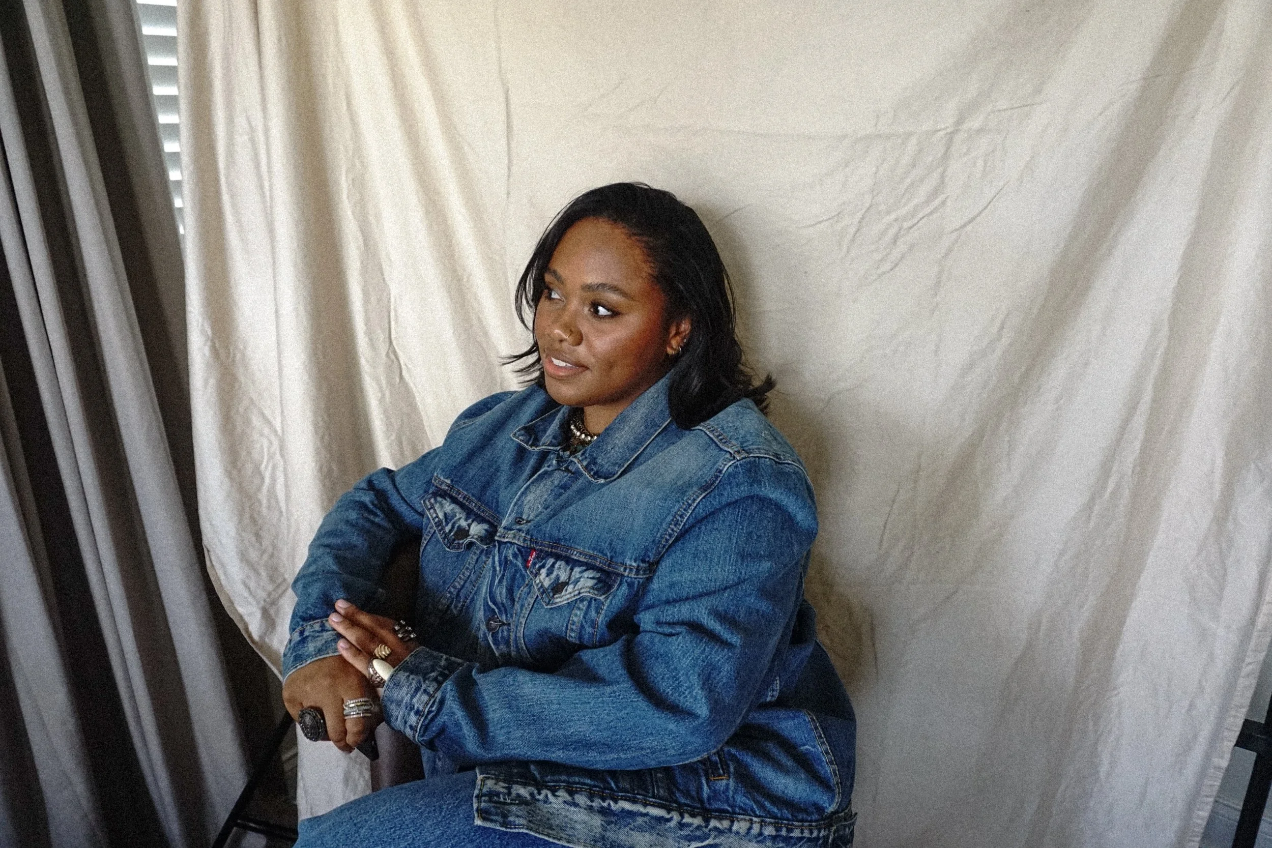 A woman with dark hair and medium skin tone sitting against a beige curtain, wearing a blue denim jacket and jeans, with jewelry including rings, a chain necklace, and earrings.
