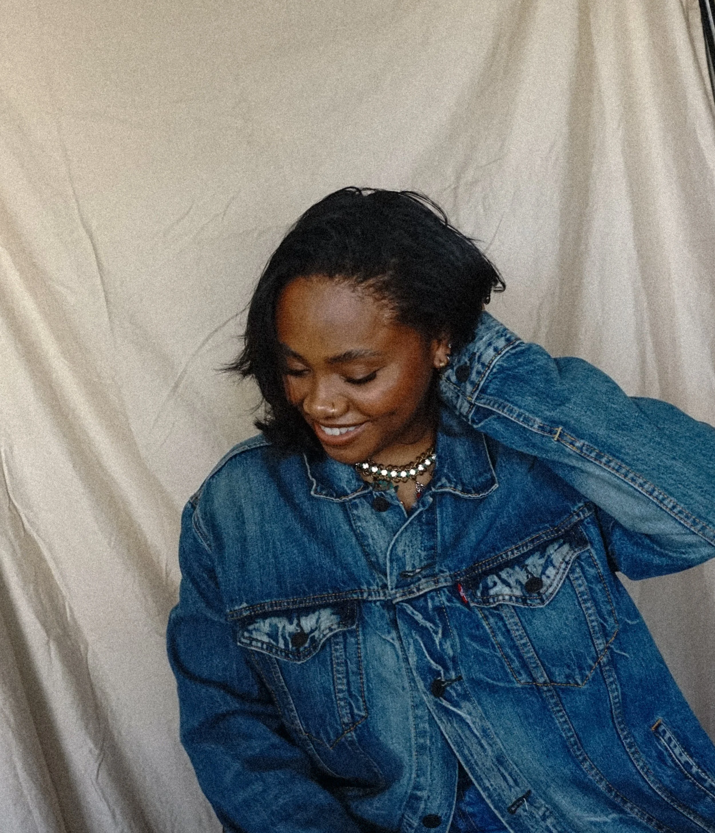 A woman with short black hair wearing a denim jacket and layered necklaces, smiling and looking down against a neutral background.