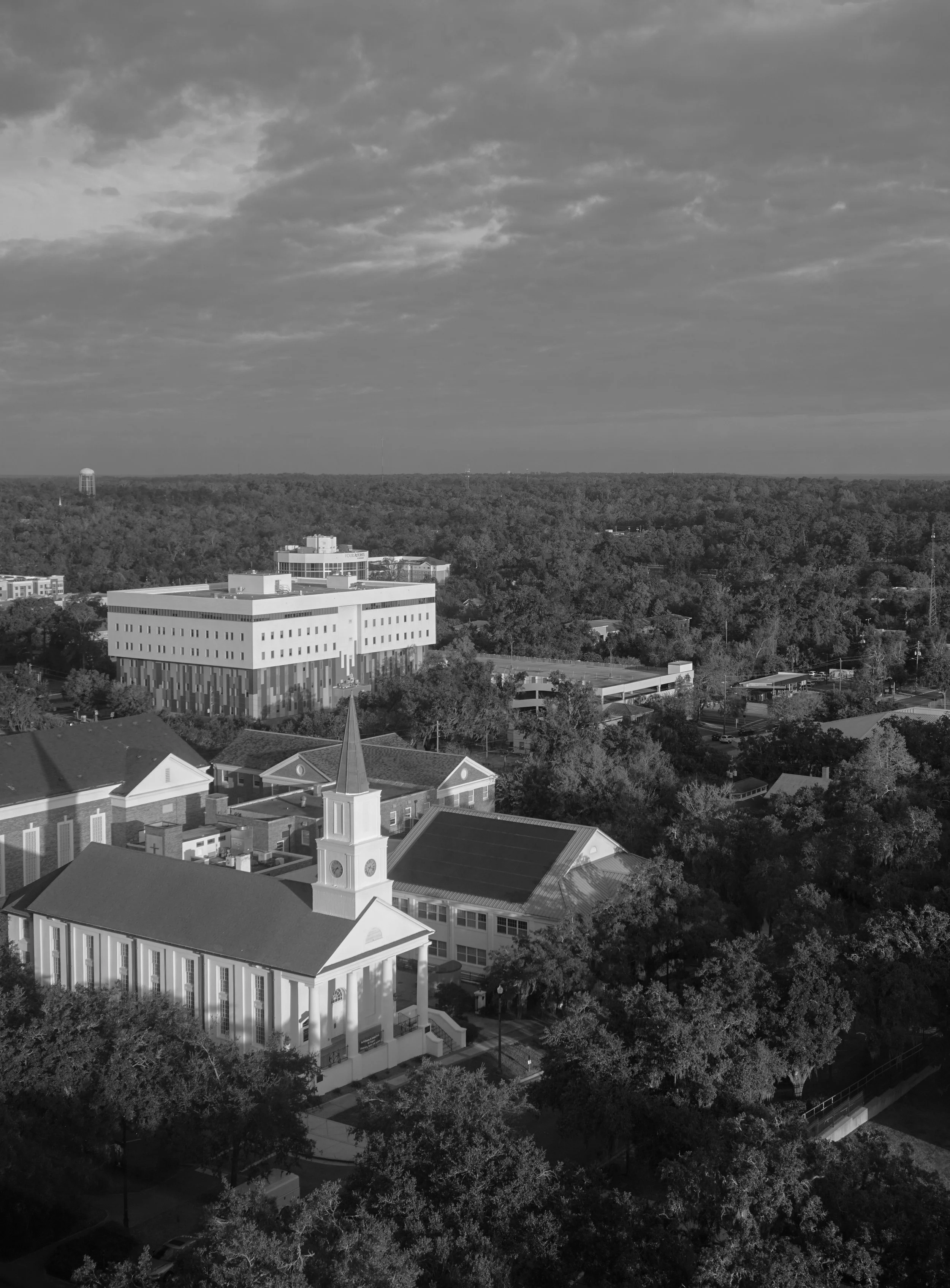 A black and white aerial view of a town with trees, a church with a steeple, and various buildings, including a modern structure in the background under a cloudy sky.