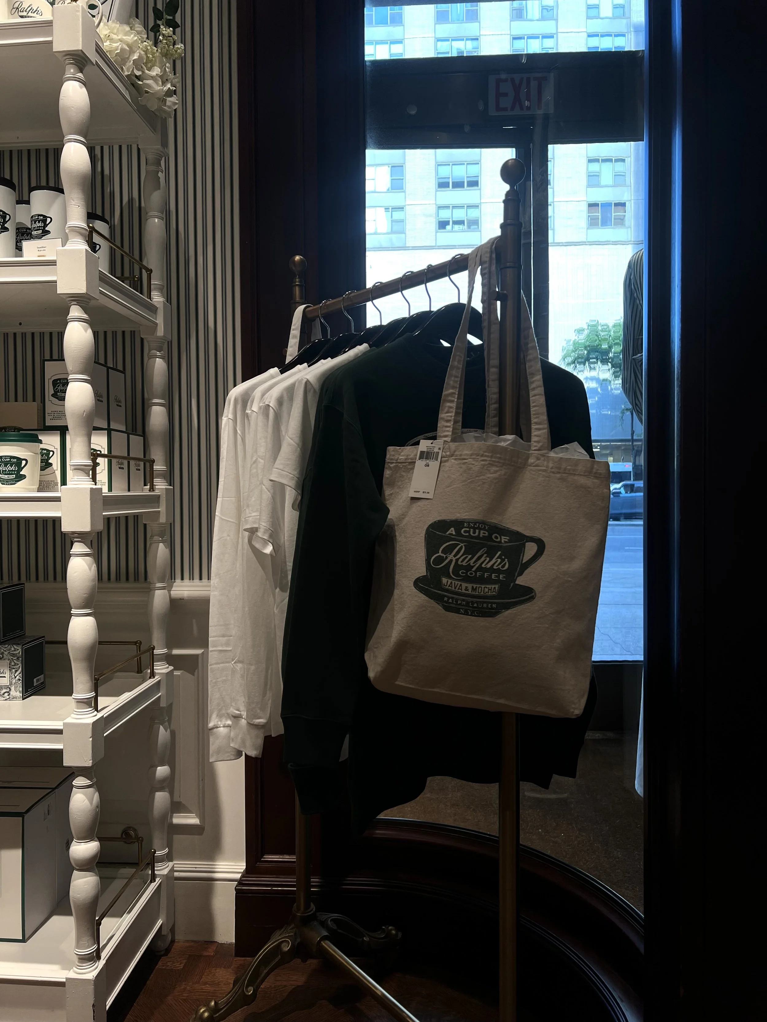 Clothing rack with white and black T-shirts and a beige tote bag with Ralph's Coffee logo in a store, with a patterned wall and a window showing city buildings in the background.