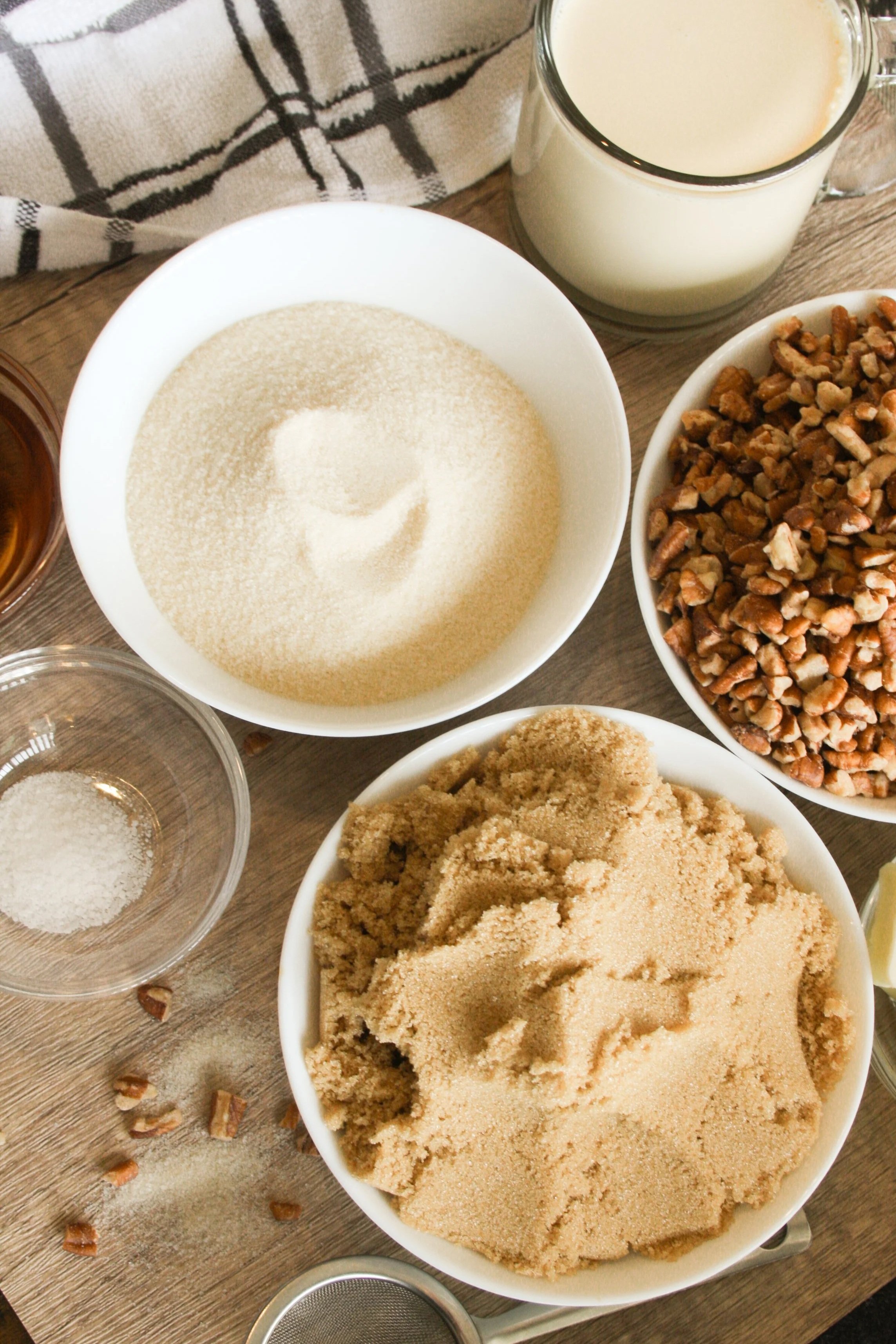 Assorted ingredients on a wooden surface, including a bowl of brown sugar, chopped nuts, powdered sugar, honey, a cup of milk, and a steel mesh strainer.