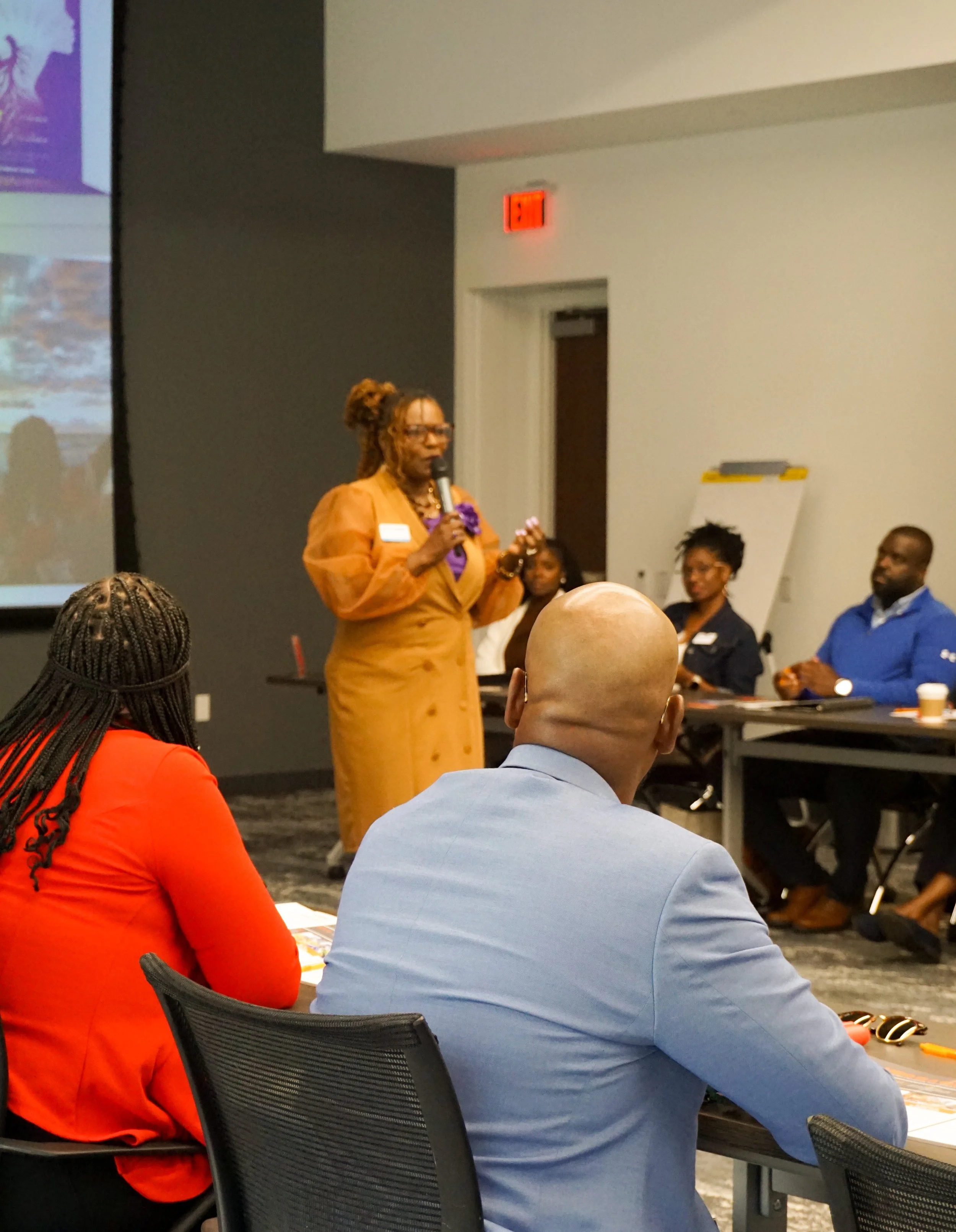 A woman in a tan dress is speaking into a microphone at a panel discussion, with four people seated at a table behind her. The audience members in the foreground are listening, with a woman in a red blazer and a man in a light blue suit visible.