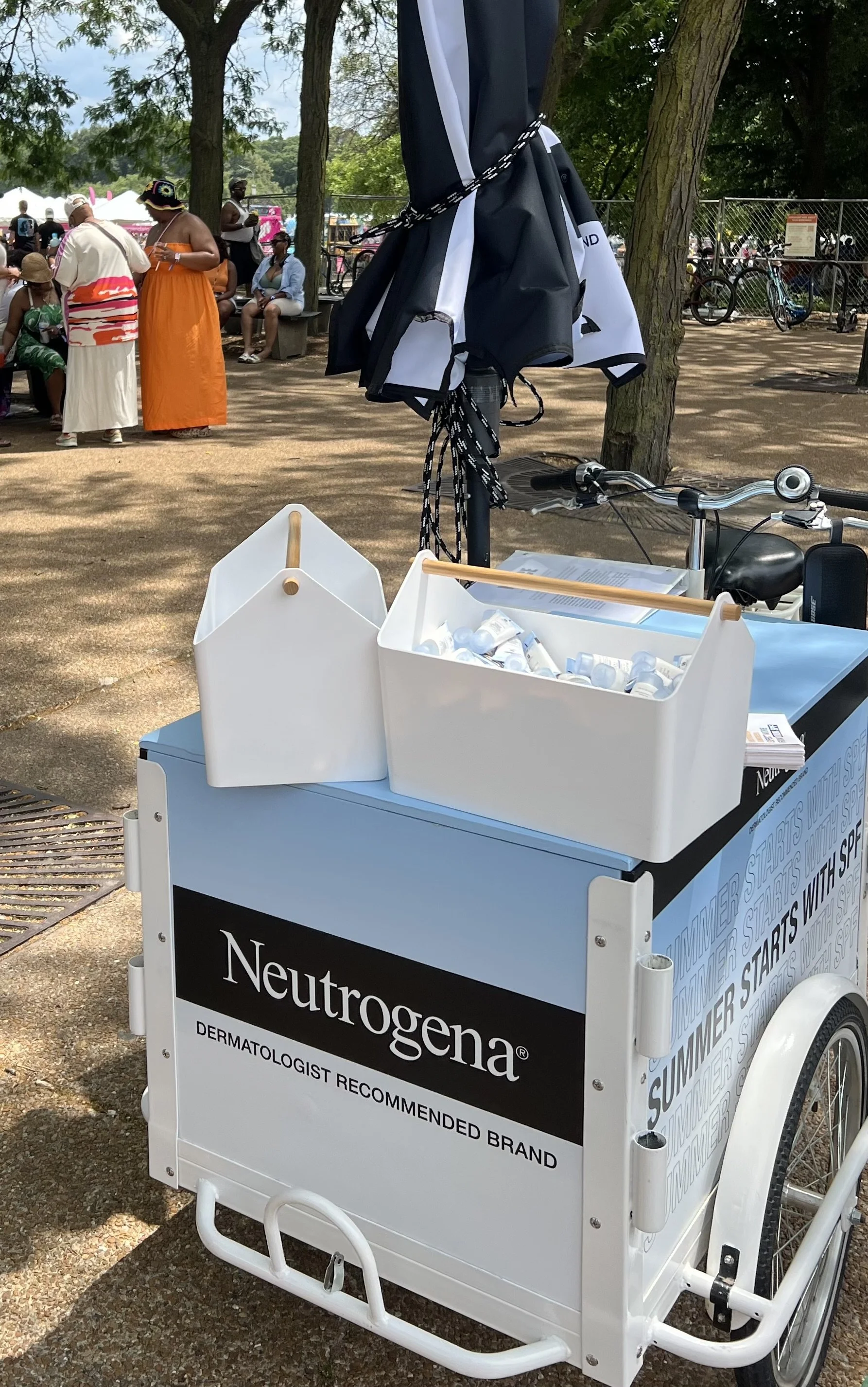 A Neutrogena skincare sample cart at an outdoor event, with sunscreen bottles in a white container on top. People are sitting and standing in the background, with some dressed in summer clothing.