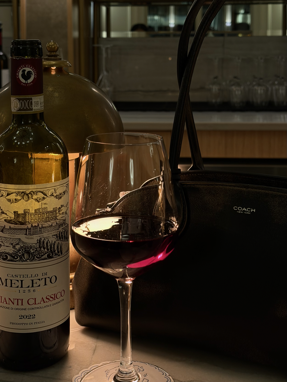 A glass of red wine on a table next to a bottle of Italian red wine, a black Coach purse, and a decorative lamp in a restaurant or cafe setting.