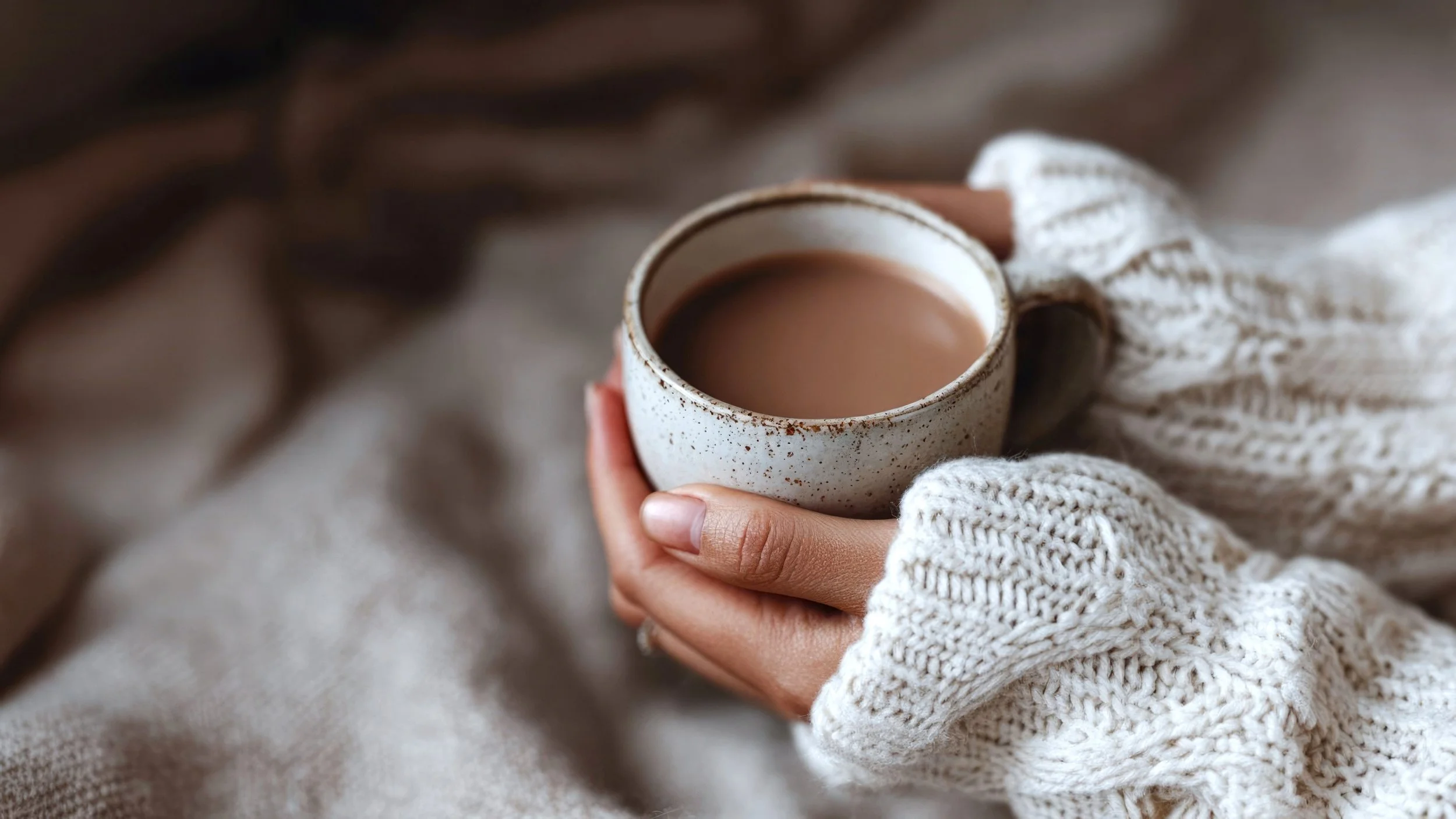 Person holding a speckled ceramic mug filled with hot chocolate or coffee, wearing cozy white knit mittens and a cream cable knit sweater, surrounded by soft blankets.