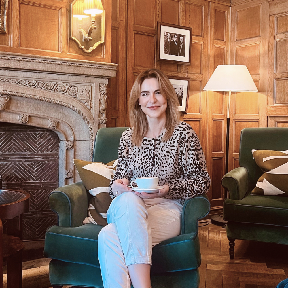 A woman with light brown hair sitting in a green armchair, holding a teacup and saucer, in a cozy wood-paneled room with framed photos on the wall and a fireplace.