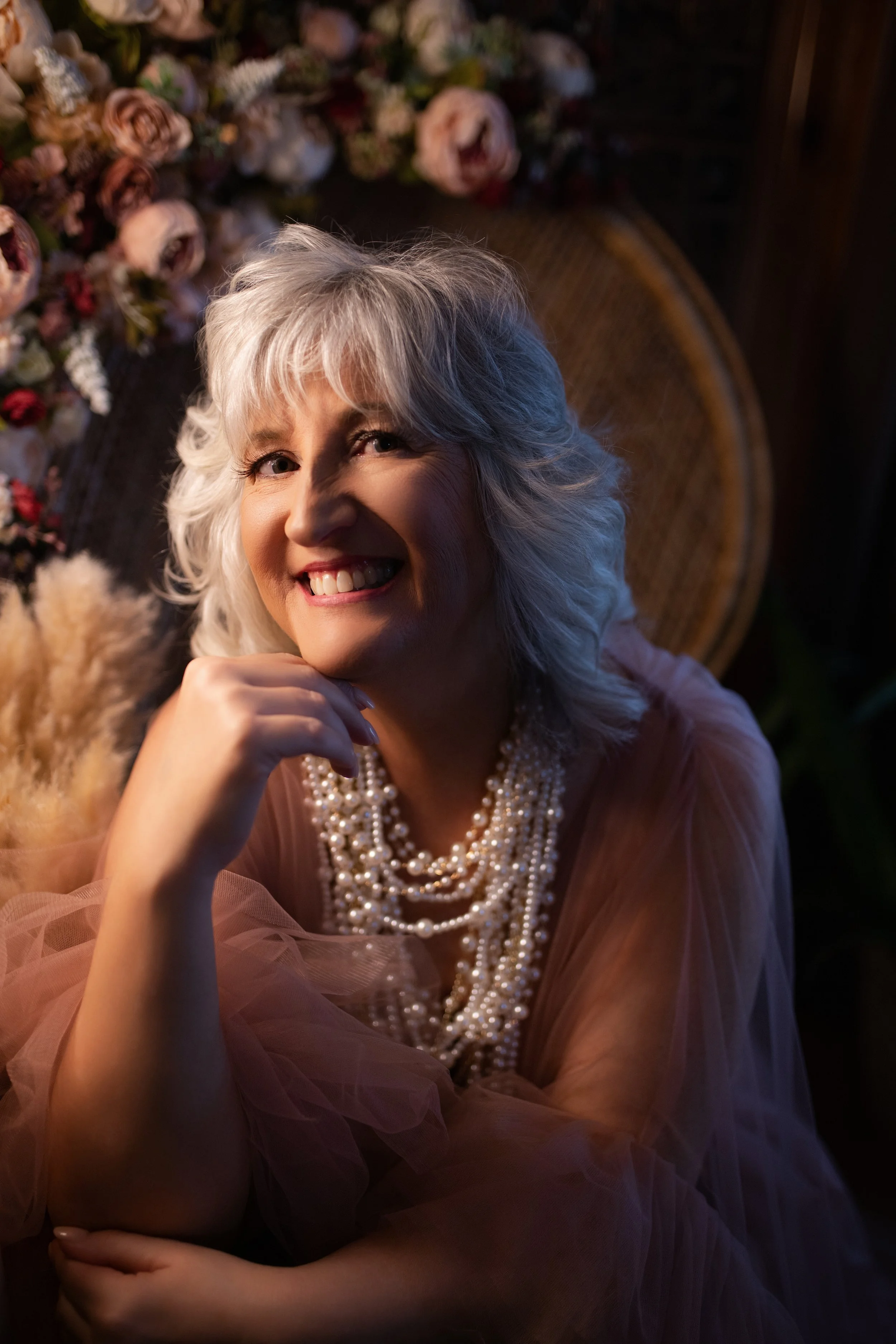 Smiling older woman with gray hair wearing a peach-colored dress and layered pearl necklaces, sitting in front of a floral backdrop and leaning on her hand.