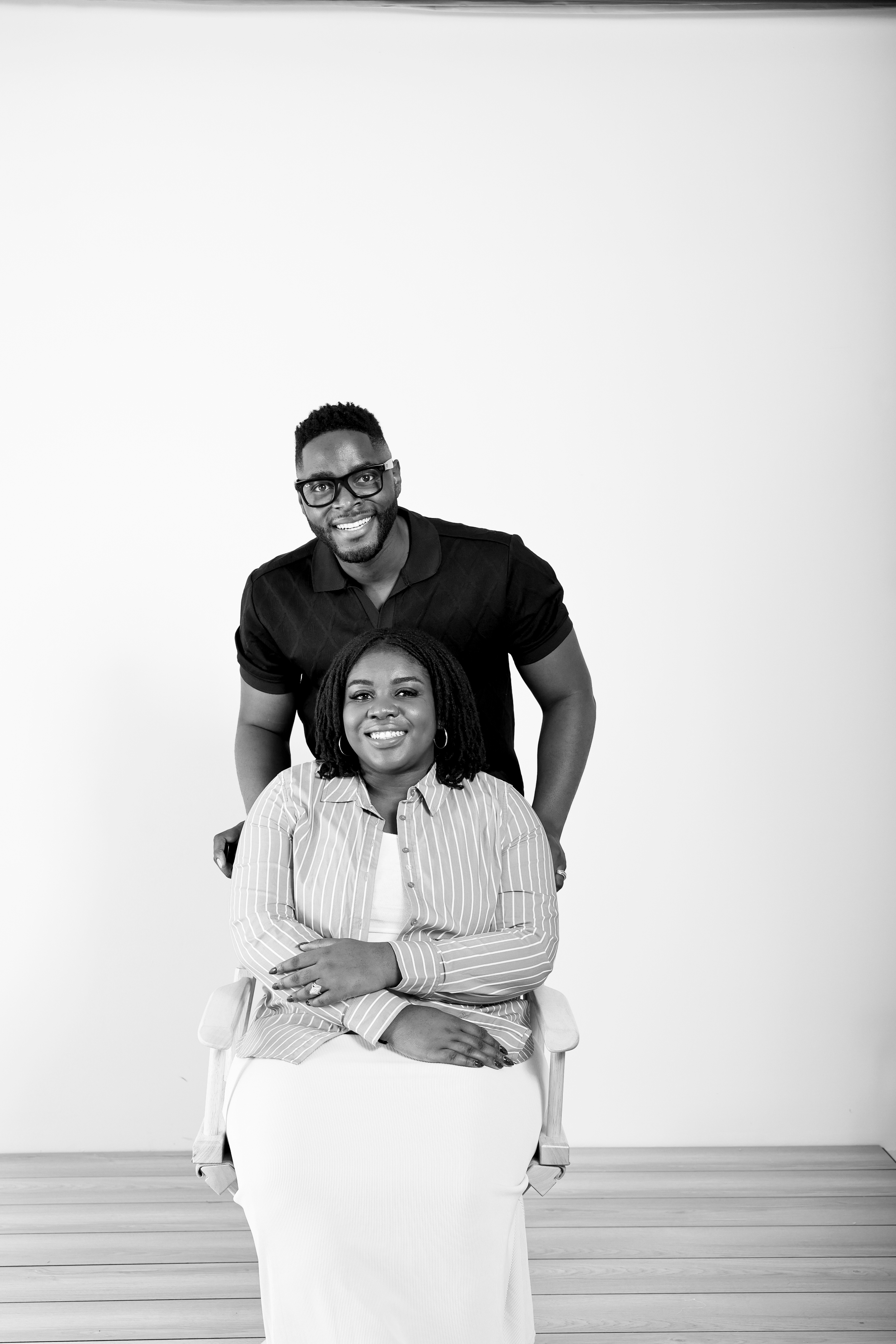 A smiling man in a black shirt stands behind a seated woman in a striped shirt and white skirt, both facing the camera, in a simple indoor setting with a plain background.
