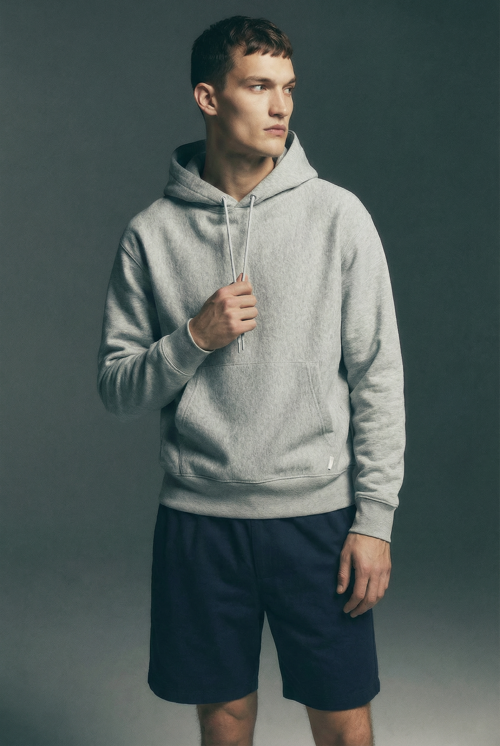 Man wearing a light gray hoodie and navy shorts standing against a plain gray background for e-commerce catalog shoot for PDP.
