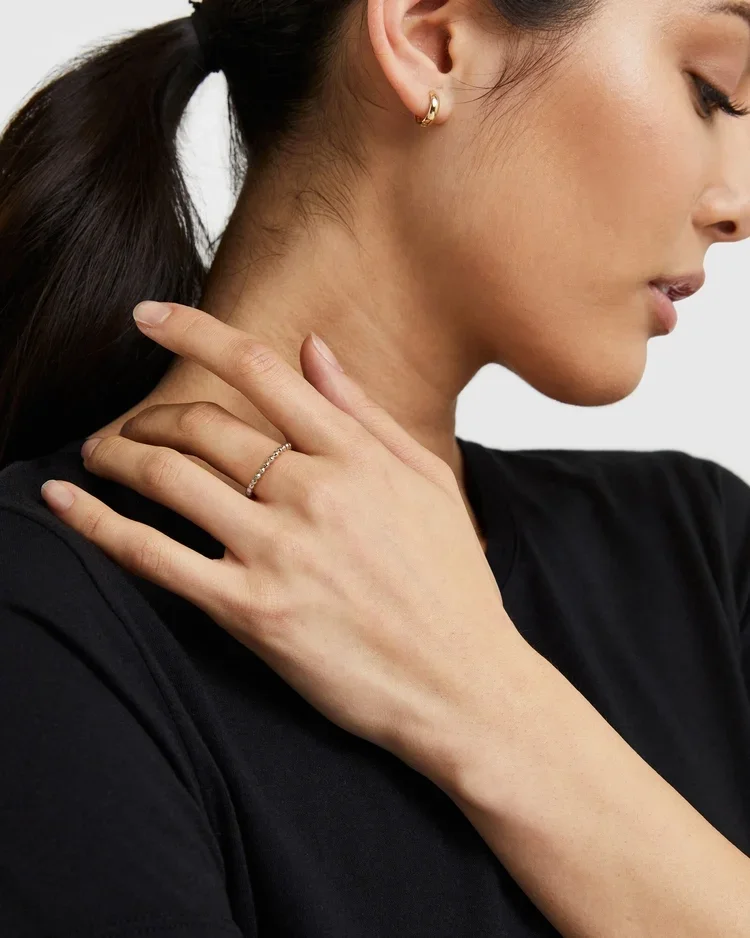 Close-up of a woman with dark hair in a ponytail wearing a black top, gold hoop earring, and a delicate ring on her finger, touching her neck with her left hand photographed for e-commerce catalog for jewelry brand PDP and emailers.