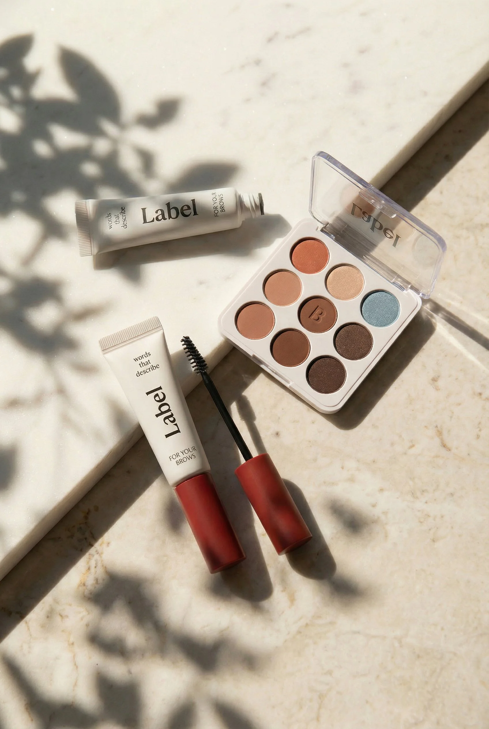An eyeshadow palette with nine shades, a brow gel, and a mascara with a curved wand on a marble surface with shadows of leaves.
