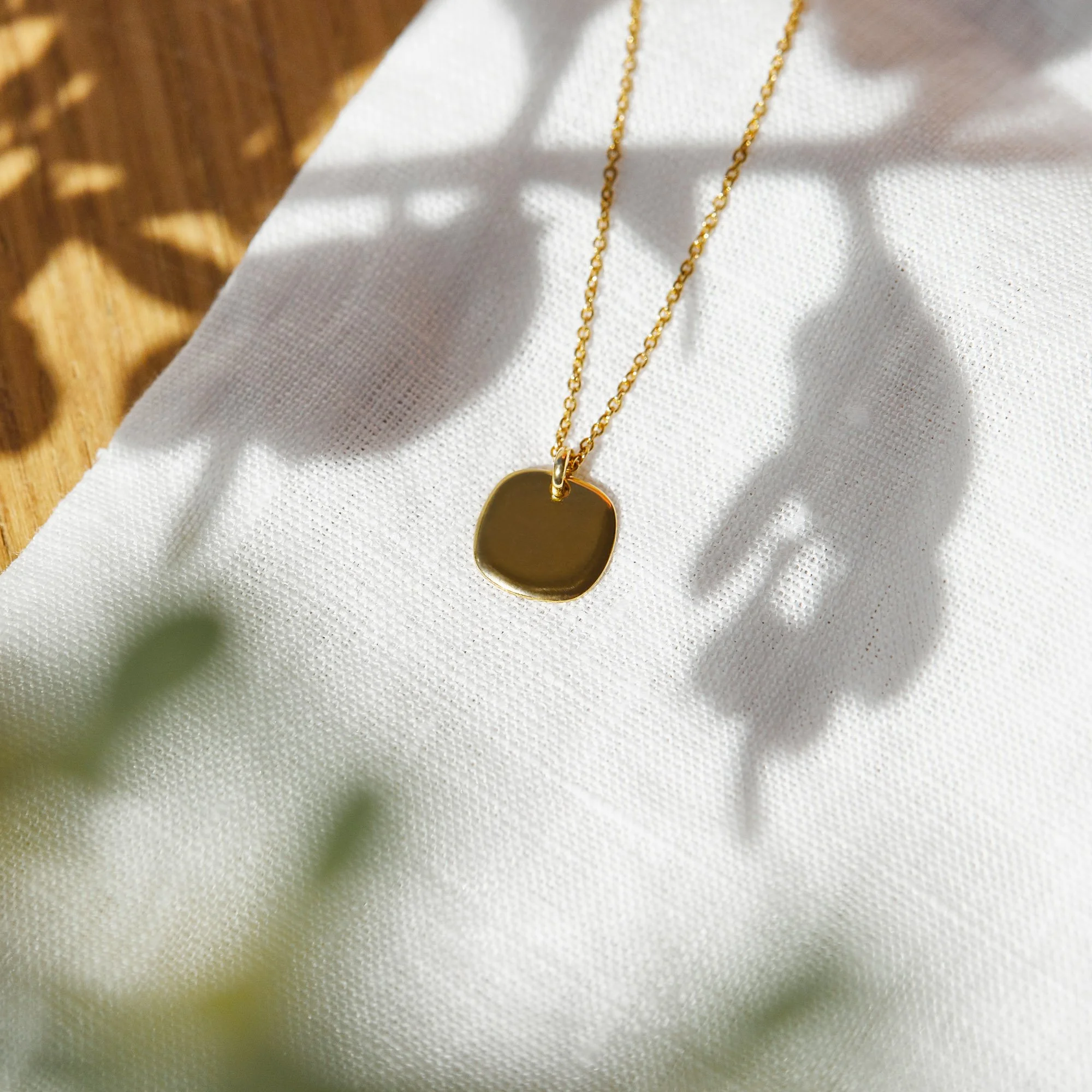 A gold necklace with a square pendant resting on a white textured fabric, casting soft shadows.