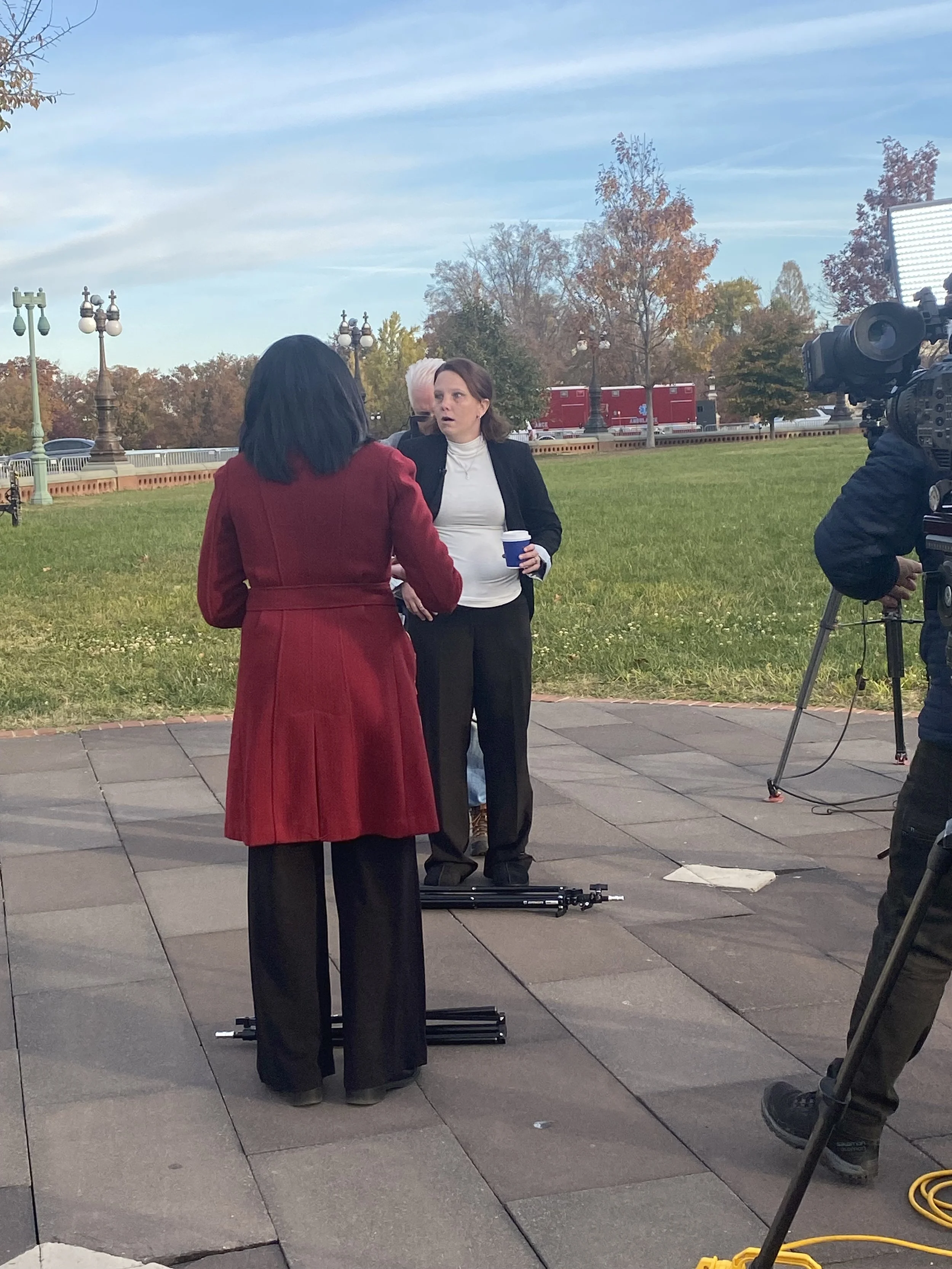 Two women are standing and talking outdoors, with a camera and a person operating it capturing the scene. One woman is wearing a red coat, and the other is holding a coffee cup and wearing a black blazer and white shirt. There are trees with fall fol