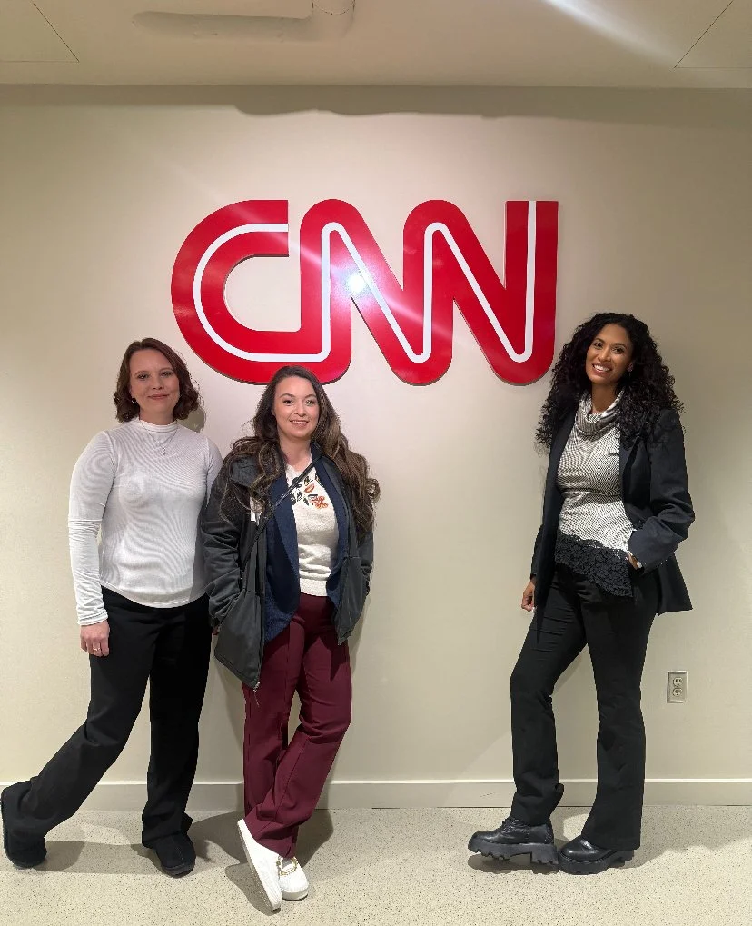 Three women standing in front of a wall with a large red CNN logo, inside an office building.
