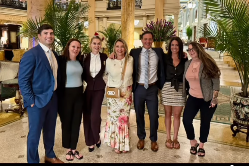 Group of seven people dressed in formal attire standing together in an ornate hotel lobby with large columns, decorative ceiling, and potted plants.