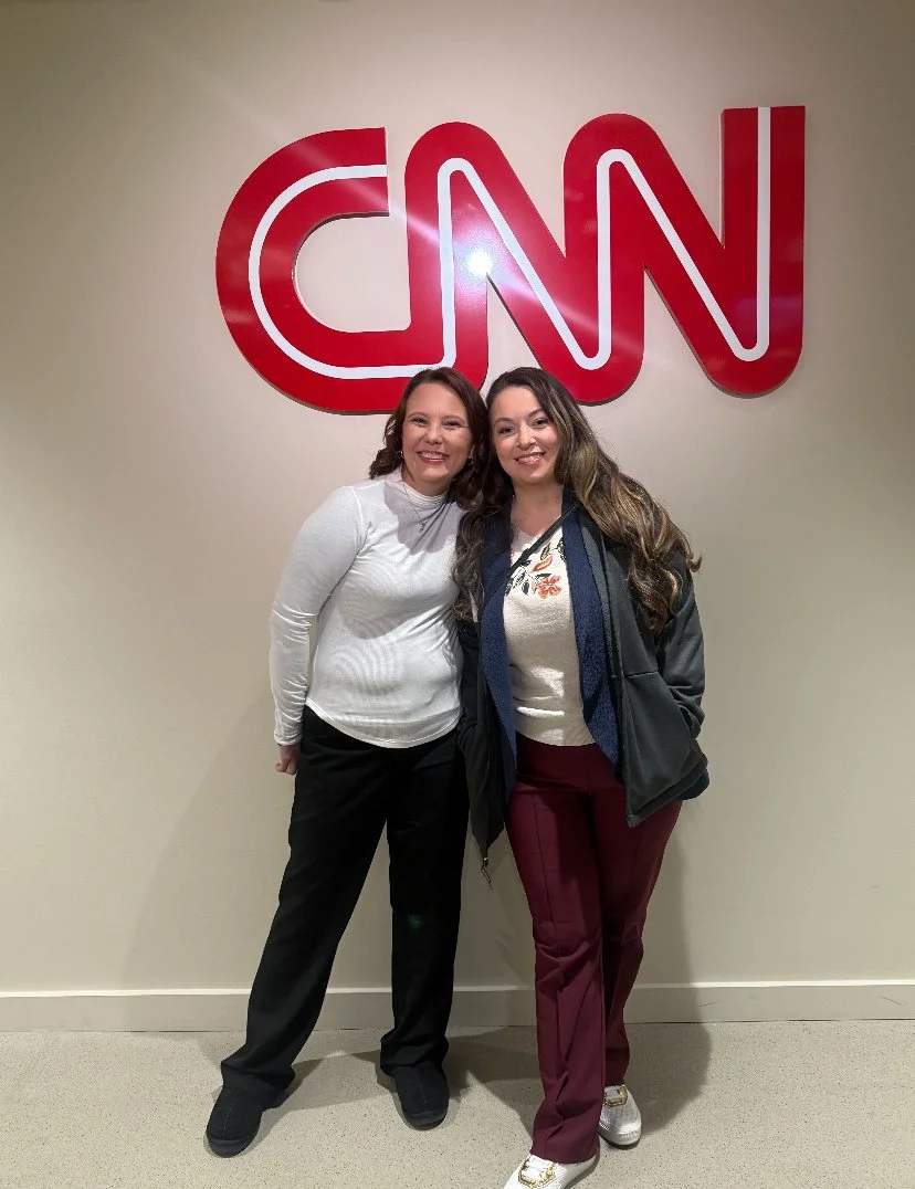 Two women standing in front of a large red CNN logo on a beige wall. They are smiling, with one wearing a white sweater and black pants, and the other wearing a patterned blouse, maroon pants, and a dark jacket.