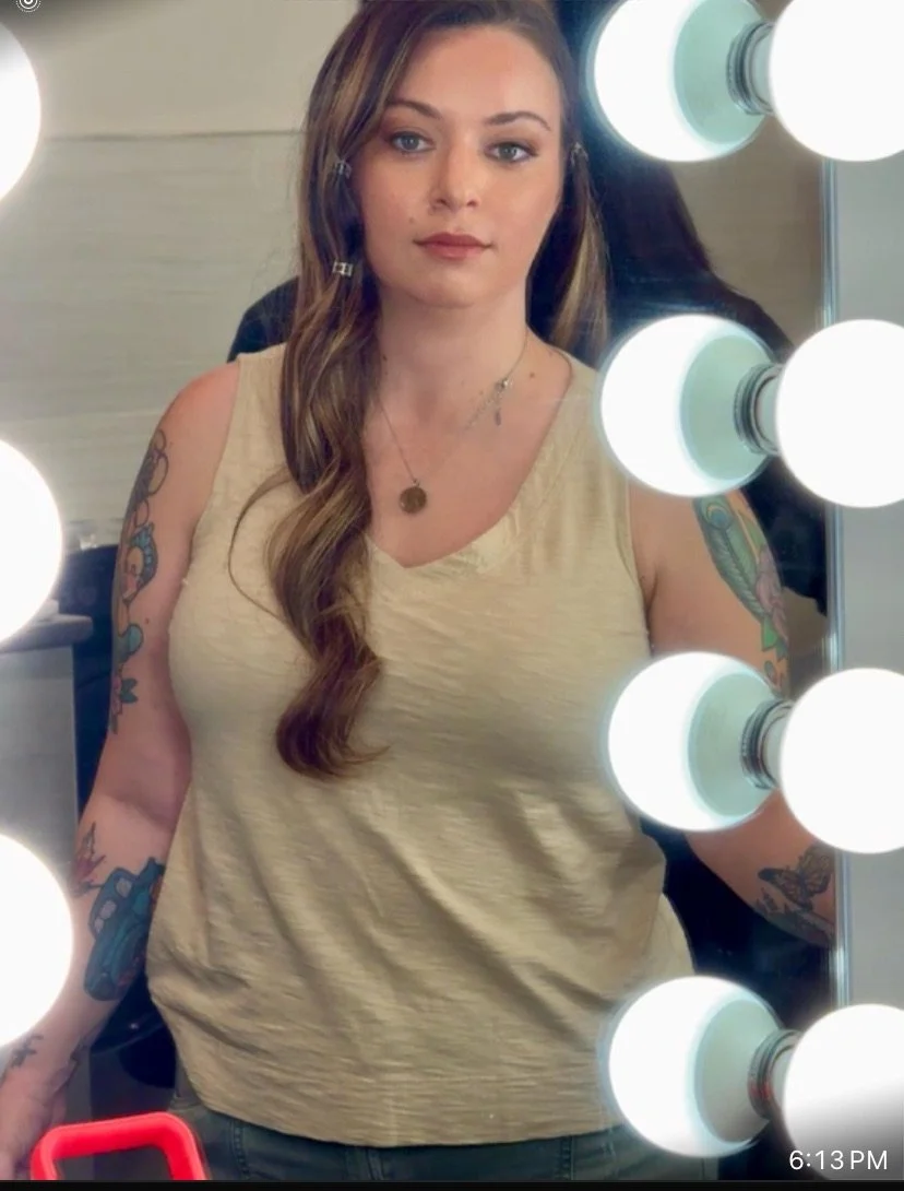 A woman taking a selfie in front of a mirror with bright vanity lights. She has long, wavy brown hair, tattoos on her arms, and is wearing a beige sleeveless top with a necklace.