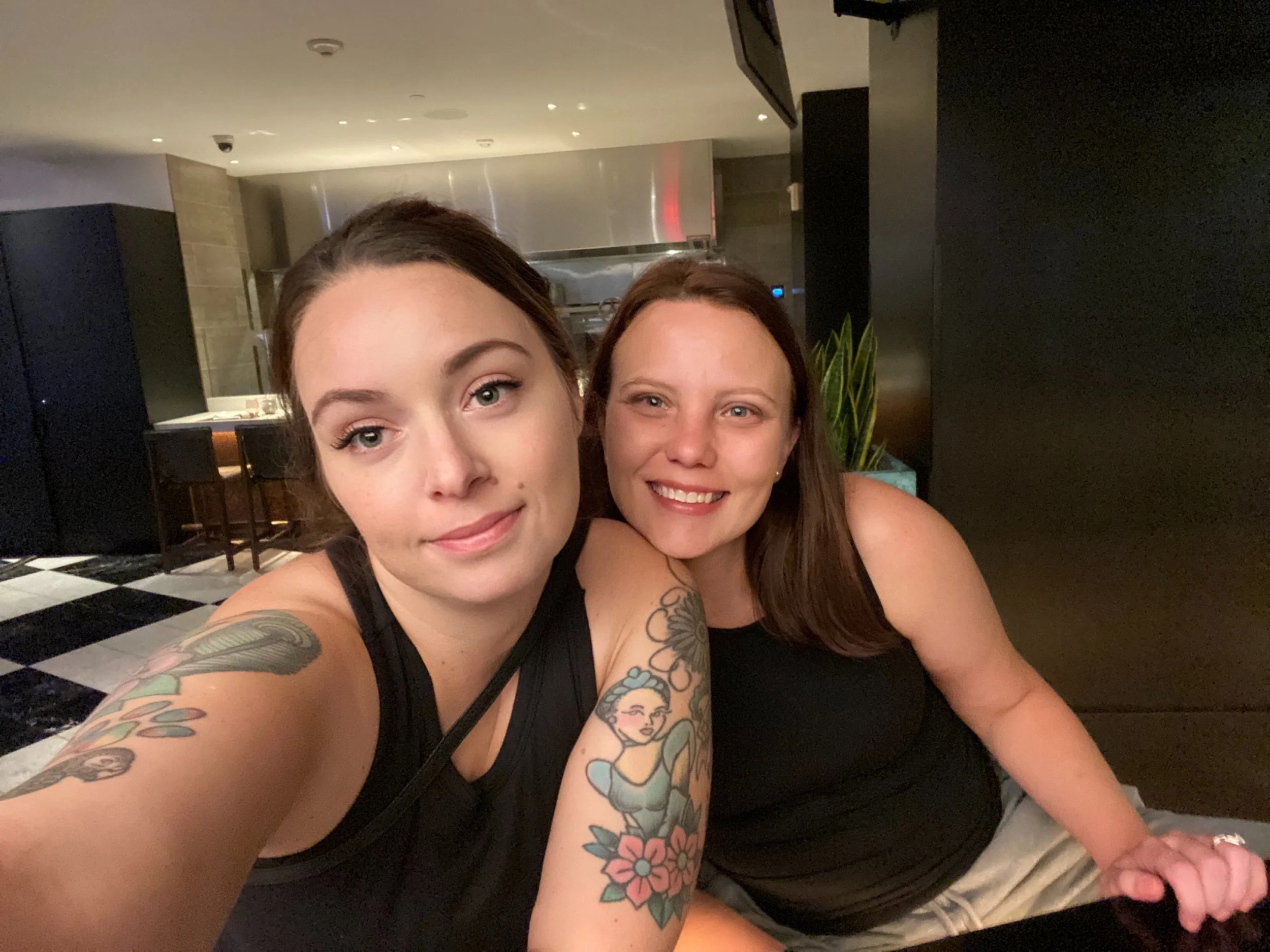 Two smiling women in black tank tops sitting close together in a dimly lit indoor setting, with one taking a selfie. The woman on the left has dark hair, tattoos on her arm, and a relaxed expression. The woman on the right has reddish-brown hair and 
