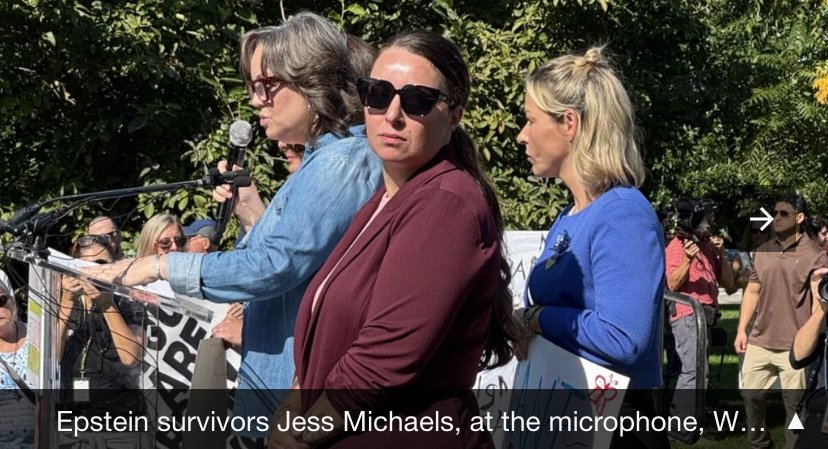 Epstein survivors Jess Michaels at the microphone during a protest or rally, surrounded by people, some holding signs, outdoors with trees in the background.