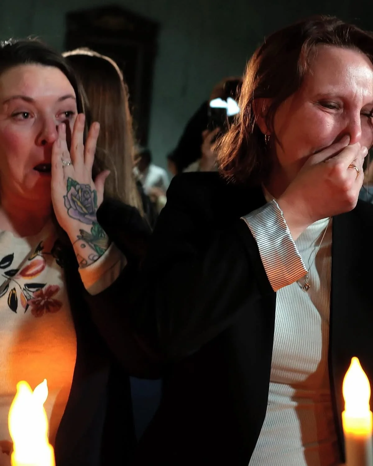 Two women at a candlelit event, one is crying and covering her mouth, the other is shocked and surprised, touching her face, both appear emotional.