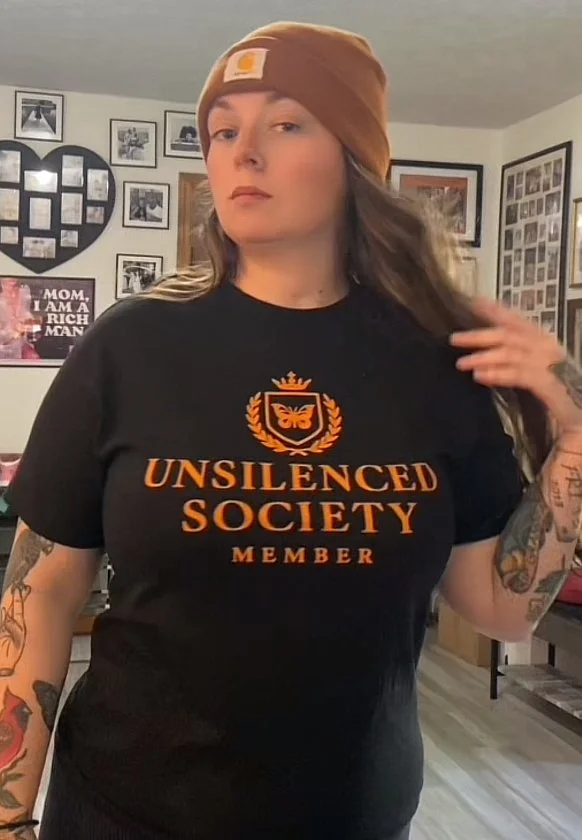 A woman wearing a brown beanie and a black T-shirt with gold lettering that reads 'Unsilenced Society Member,' standing in a room with framed photos and artwork on the walls.