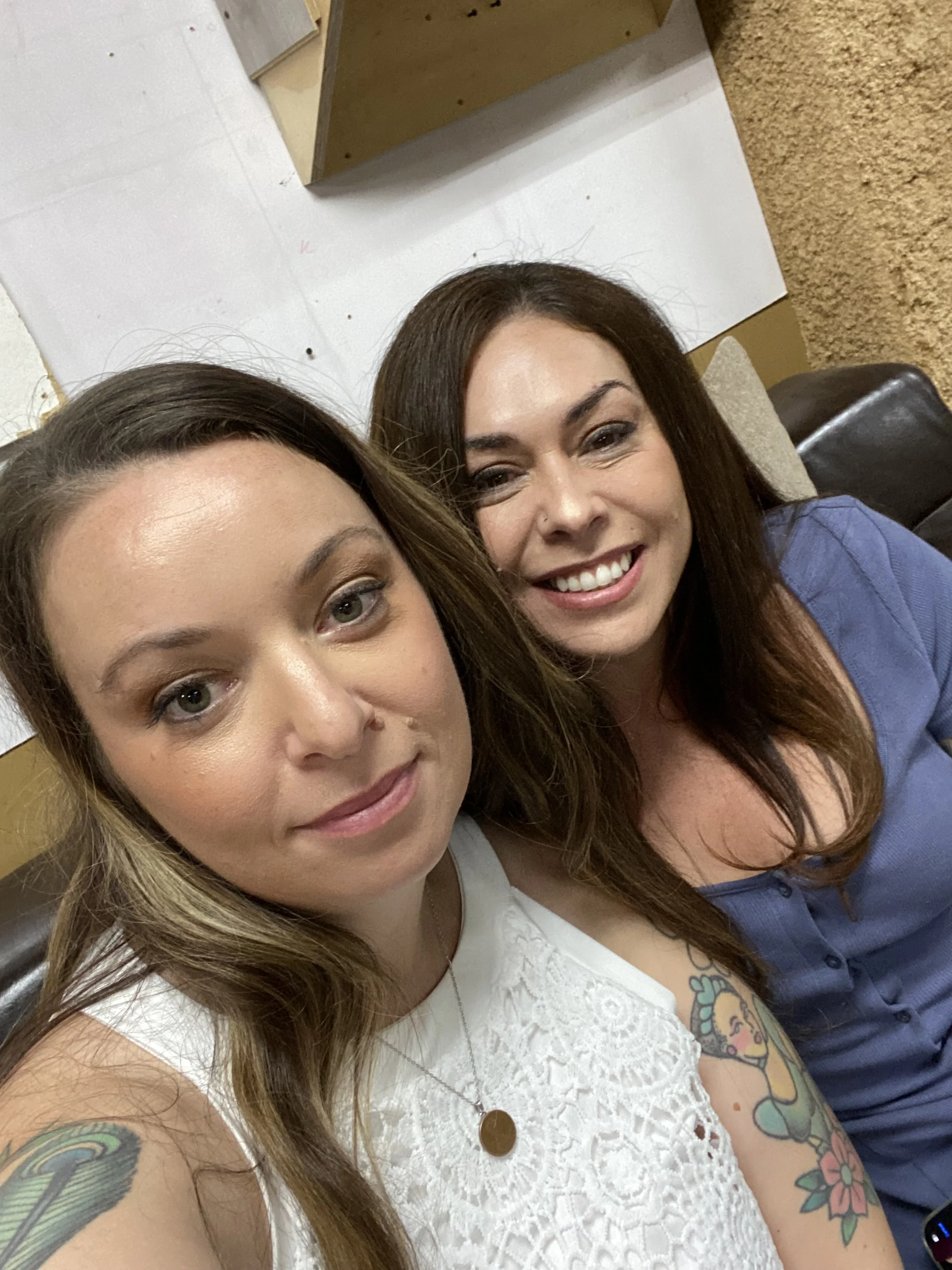 Two women taking a selfie together indoors, smiling at the camera. One woman has light brown hair, a tattoo on her upper arm, and is wearing a white lace top. The other woman has darker brown hair and is wearing a blue shirt.