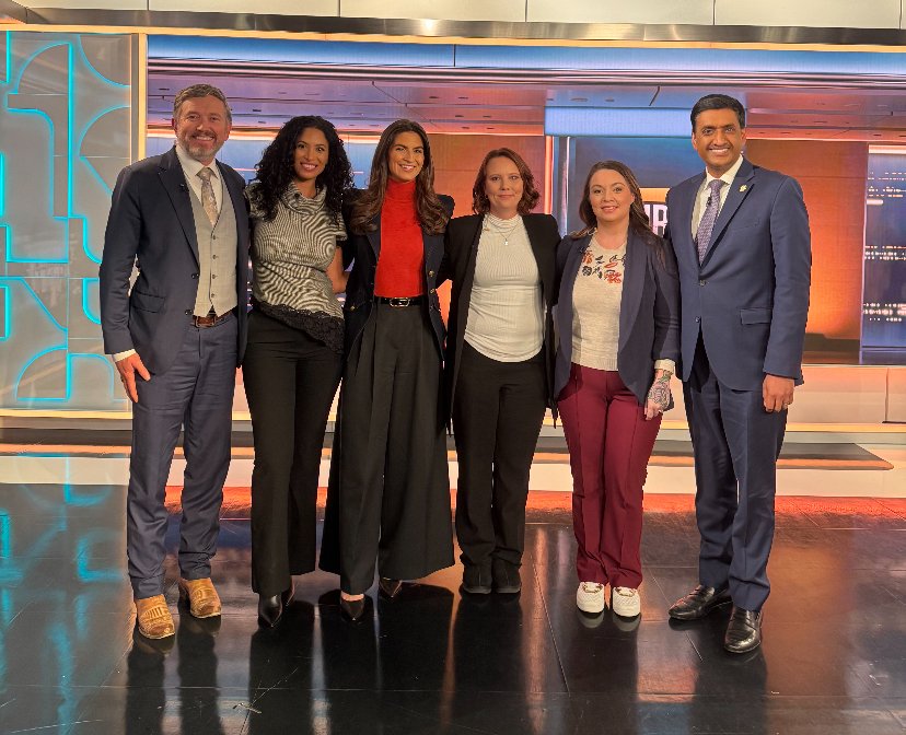 Six news anchors standing together on a TV set, smiling at the camera.