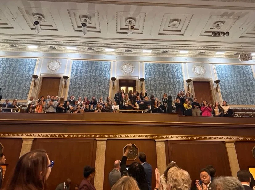 People standing and sitting in a gallery section of a large, ornate room with blue wallpaper, decorative molding, and a high ceiling with recessed lighting.