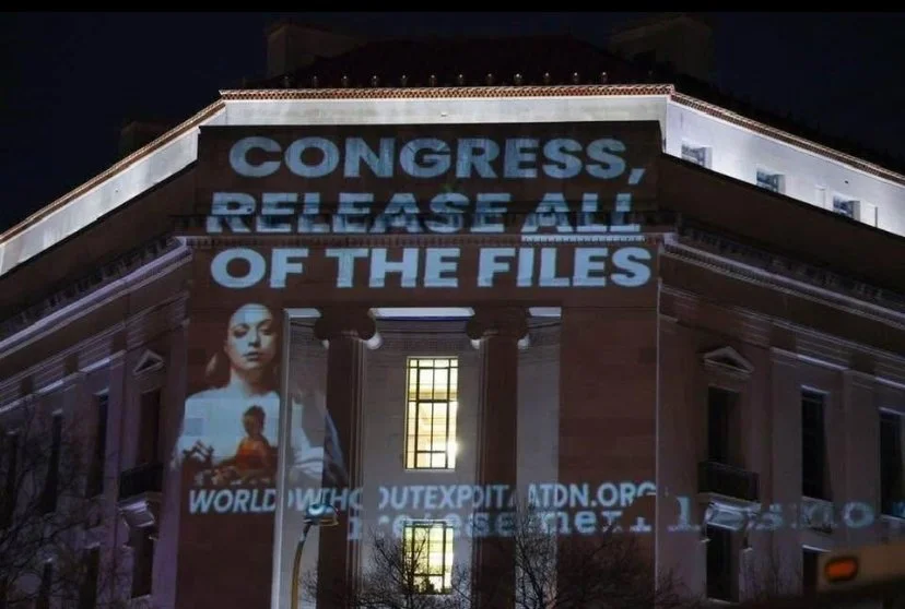 Light projection on a building displays the message: 'CONGRESS, RELEASE ALL OF THE FILES' along with images of a woman and a child, and web addresses.
