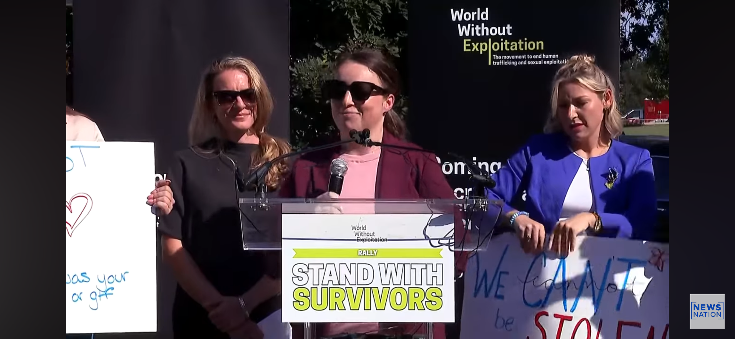Women speaking at a rally with signs; one says 'Stand with Survivors,' others support ending exploitation and stopping trafficking.