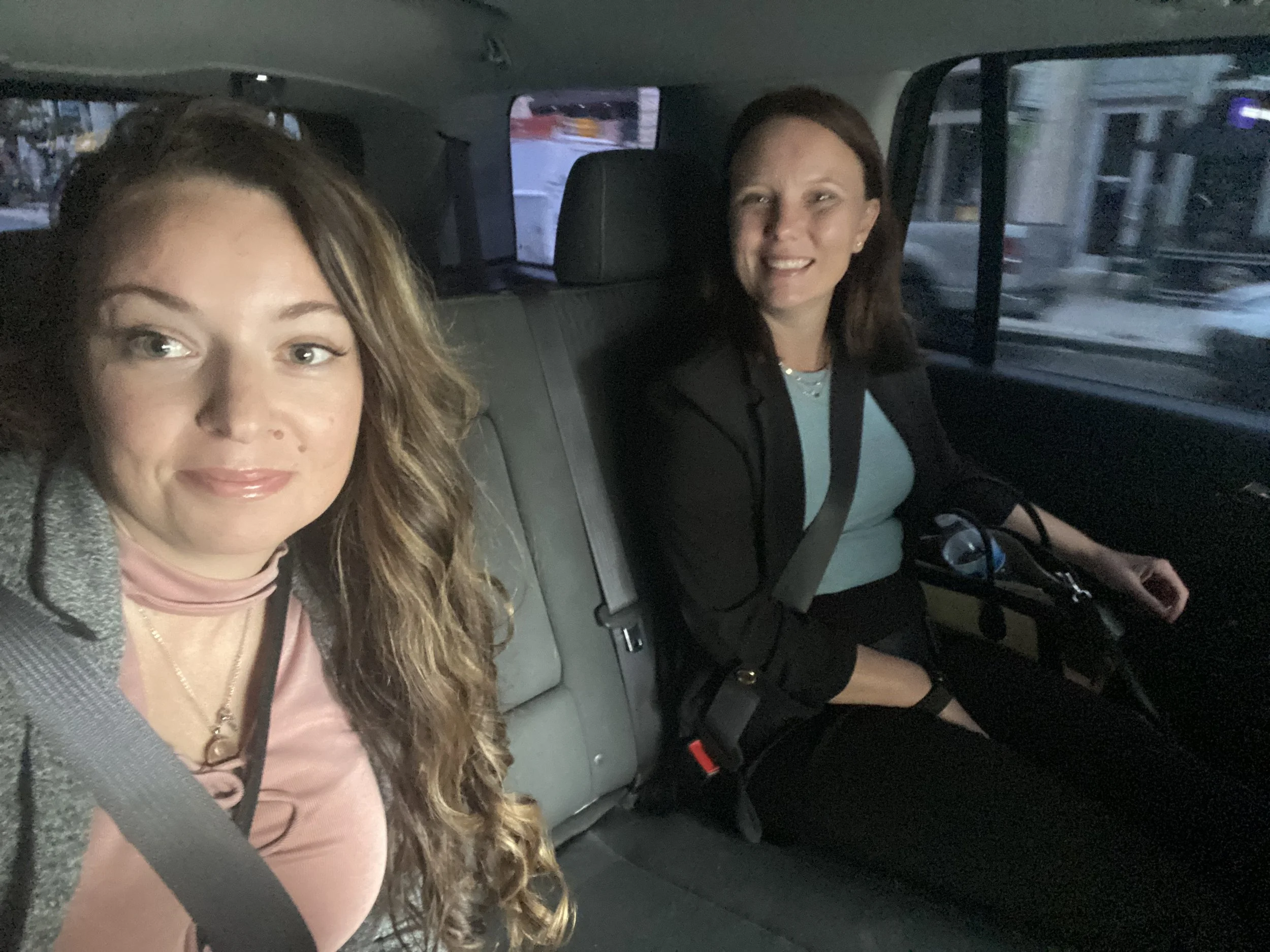 Two women sitting in the backseat of a vehicle, one taking a selfie. The woman on the left has long wavy brown hair, blue eyes, and is smiling. She wears a pink top with a gray jacket and a seatbelt. The woman on the right has shoulder-length brown h