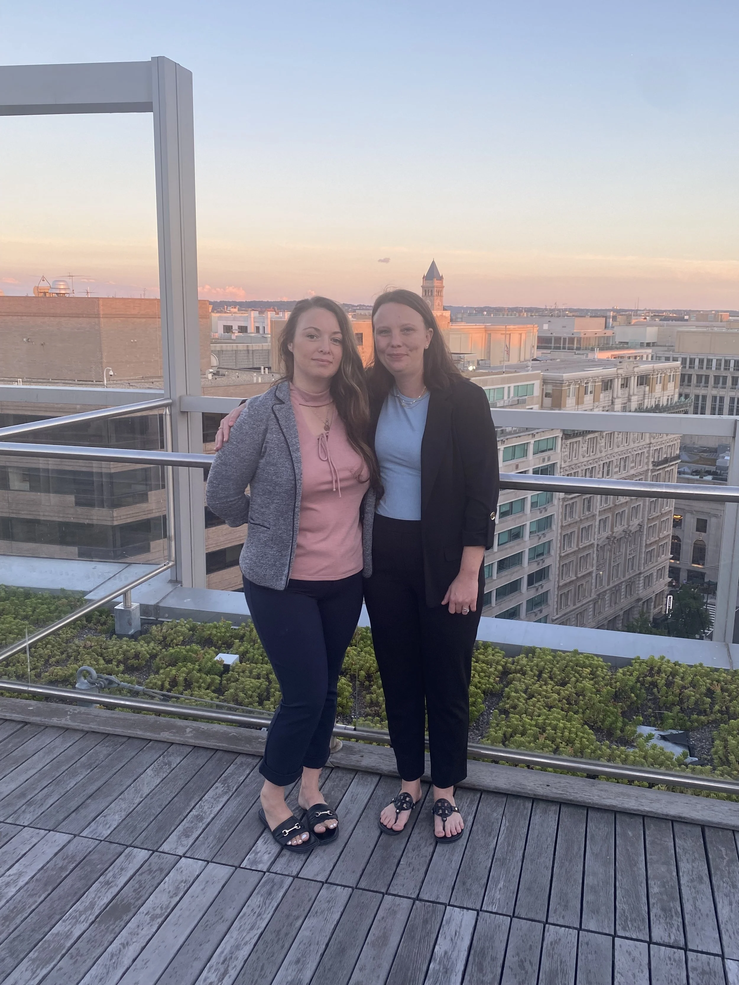 Two women standing close together on a balcony with city buildings in the background during sunset.