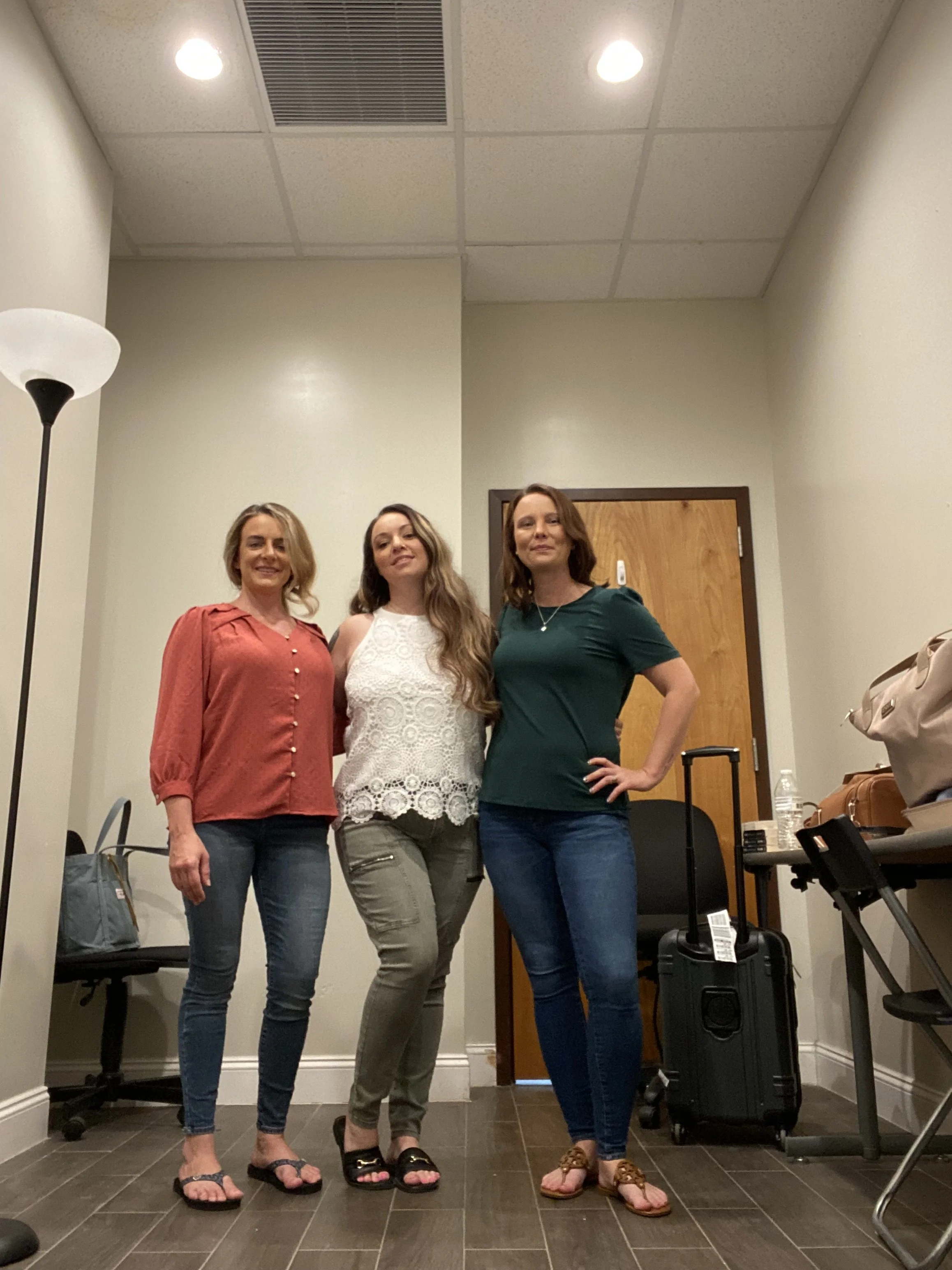 Three women standing indoors on a tiled floor, smiling at the camera, with a table, a black suitcase, a water bottle, and a beige purse on it in the background.