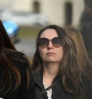 A woman with wavy brown hair wearing large sunglasses and a black jacket, walking outdoors.