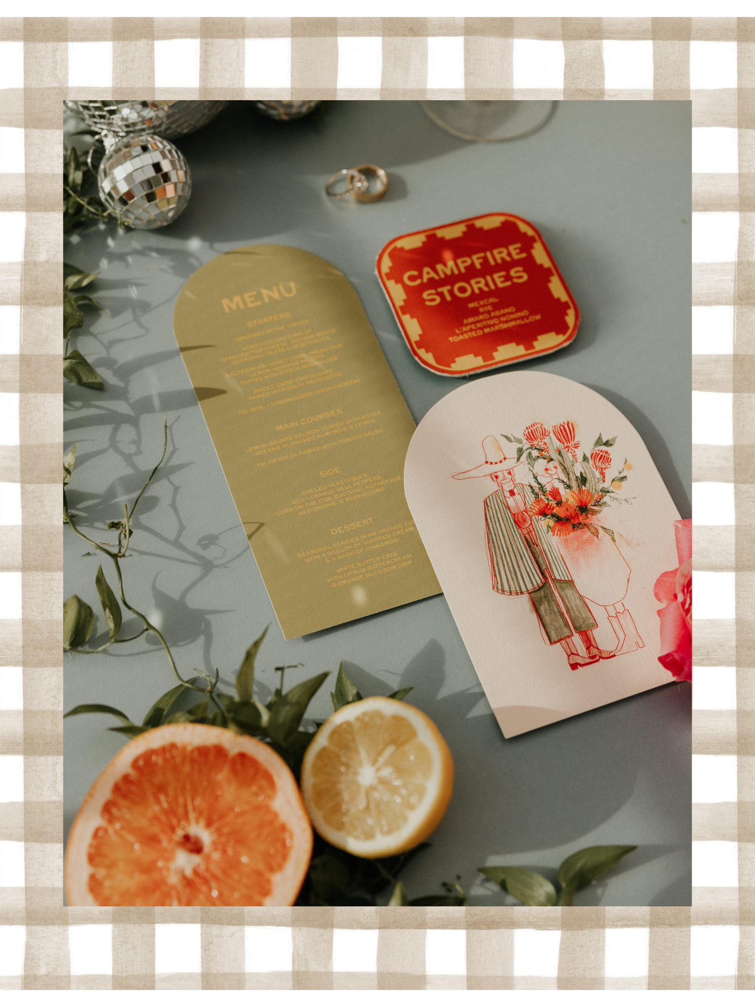 A table with two halved fruits, a grapefruit and lemon, along with a menu, a card illustration of a person with flowers, a red tin box labeled 'Campfire Stories,' rings, and decorative silver balls, over a checked beige and white border.