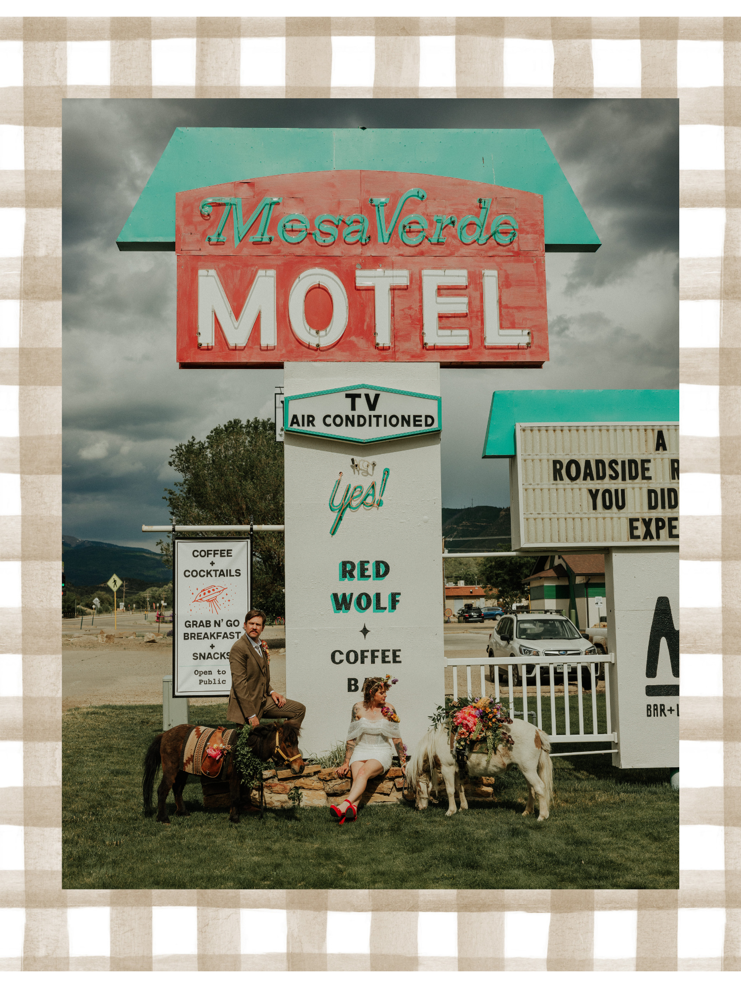 Man and woman with two small ponies in front of a vintage motel sign, with cloudy sky in the background. The man is standing with one foot on a small rock, the woman is sitting on the ground, both are dressed in vintage style.