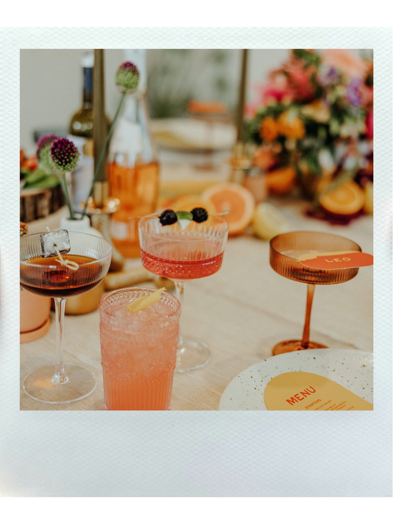 Assorted colorful cocktails and drinks on a table with flowers and a menu, with a floral arrangement in the background.