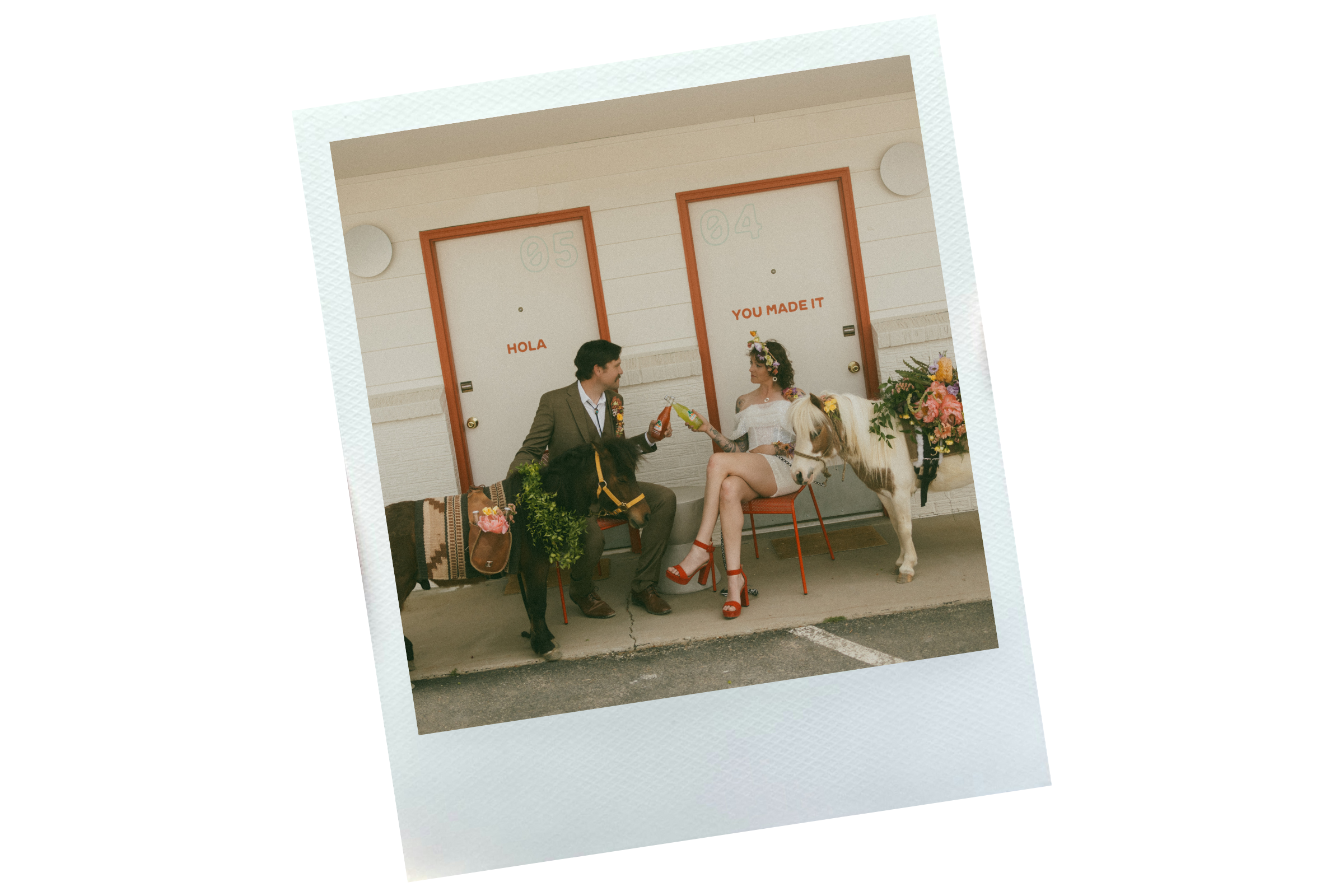 A man and woman sitting outside in front of two doors, one labeled '05 HOLA' and the other '04 YOU MADE IT.' The man is holding a drink, and the woman is wearing a floral crown and red shoes, with a small horse beside her decorated with flowers.