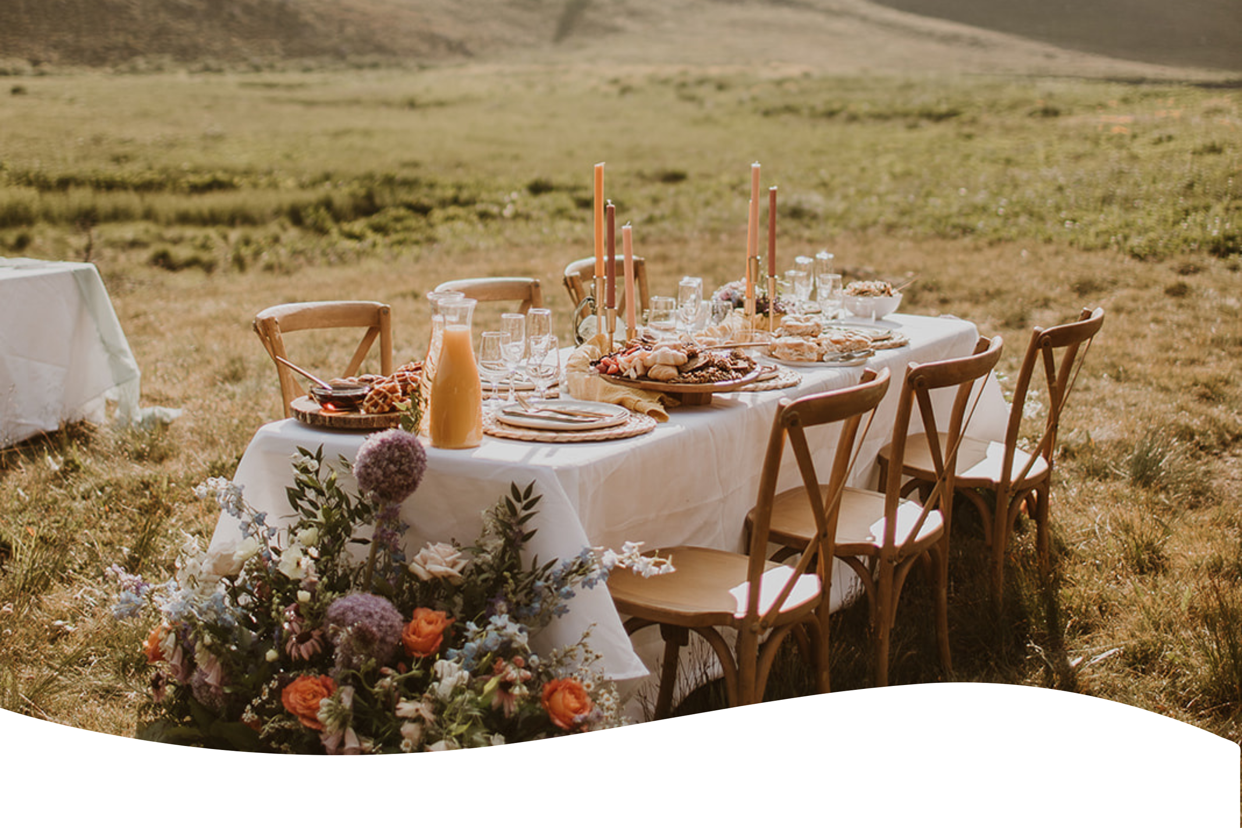 A banquet table set outdoors in a grassy field, decorated with flowers, candles, and place settings, with a background of open land.
