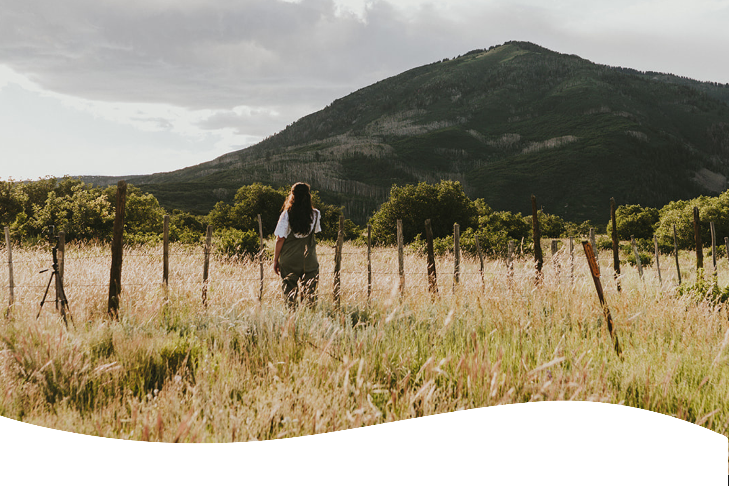 A woman with long hair walking through a grassy field with a mountain in the background, under a cloudy sky.