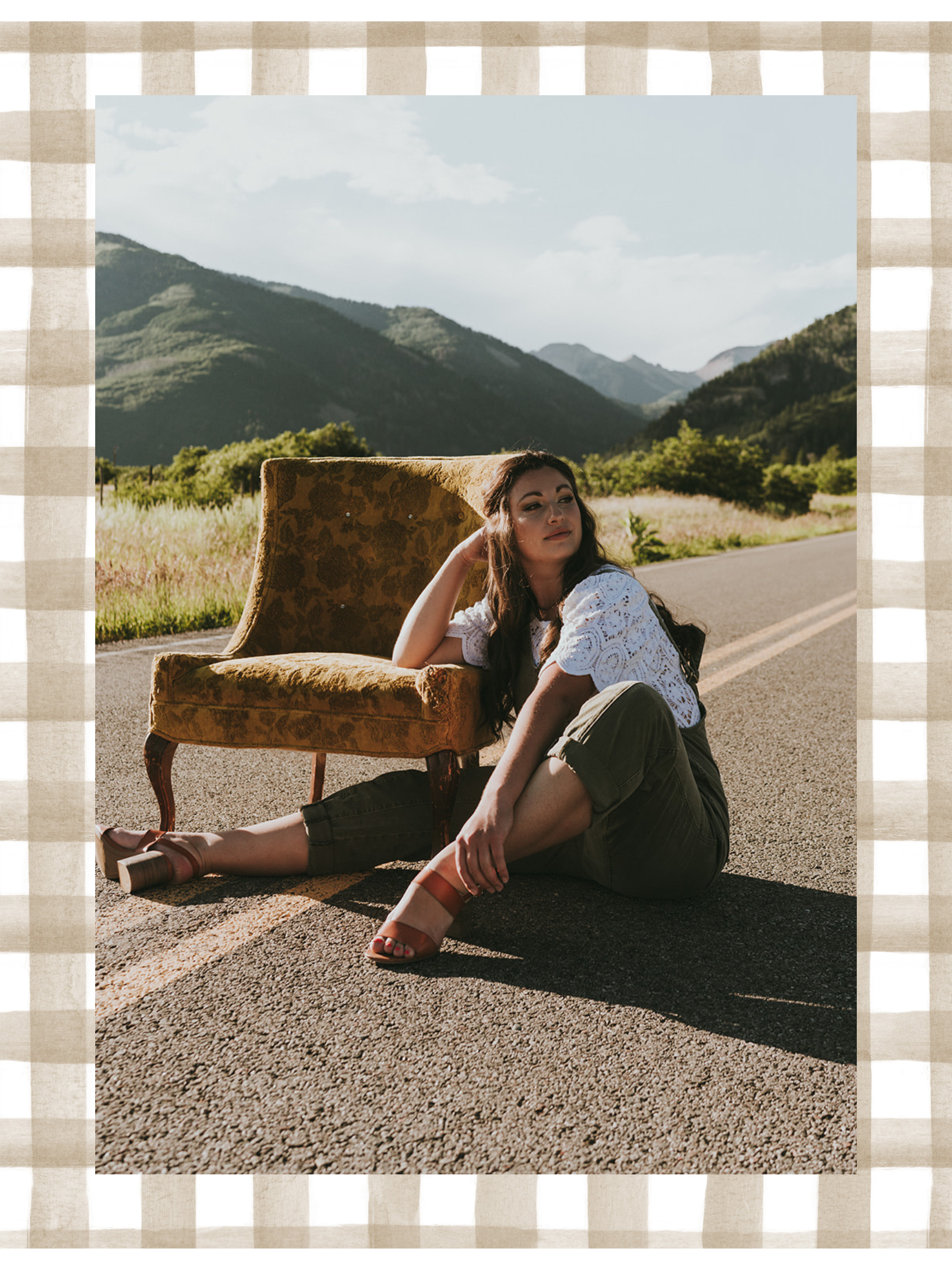A woman sits on the road next to an old sofa in a rural landscape with mountains in the background.