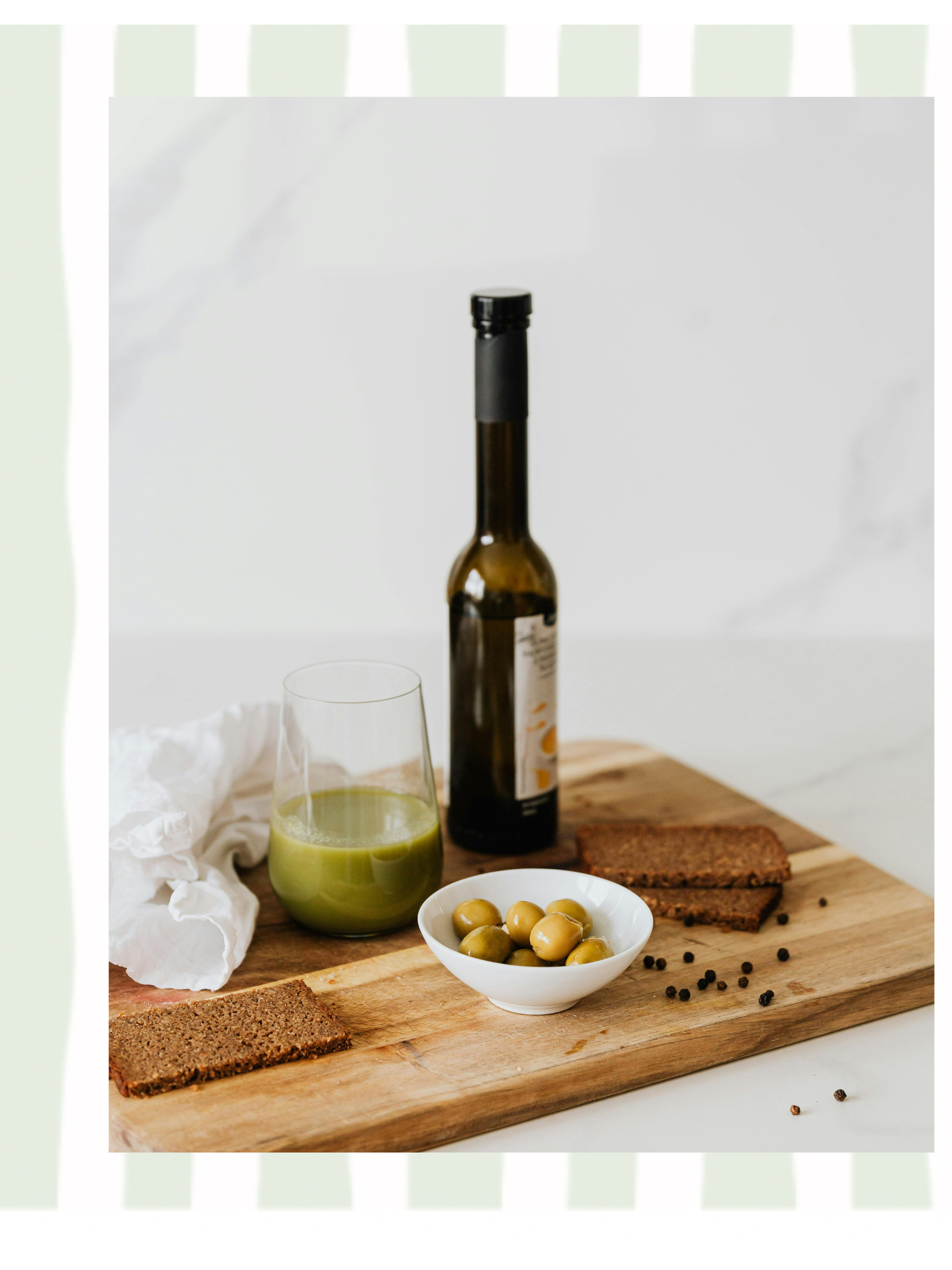 A wooden board holding a glass of green juice, a bowl of green olives, two slices of brown bread, a bottle of olive oil, and scattered black peppercorns, with a white cloth and a white marble background.