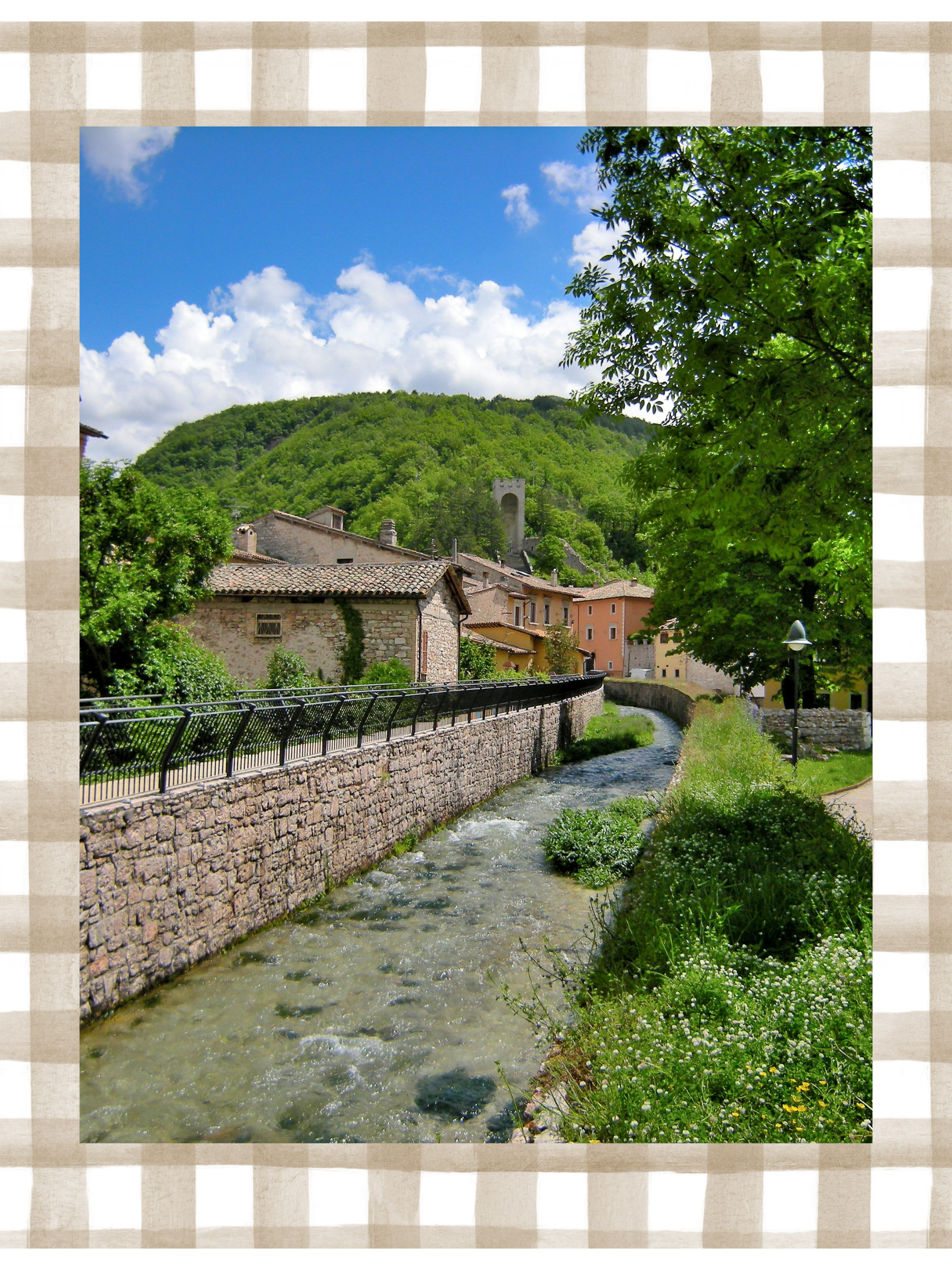 A picturesque village with colorful buildings along a river, surrounded by green trees and hills, with a partly cloudy blue sky.