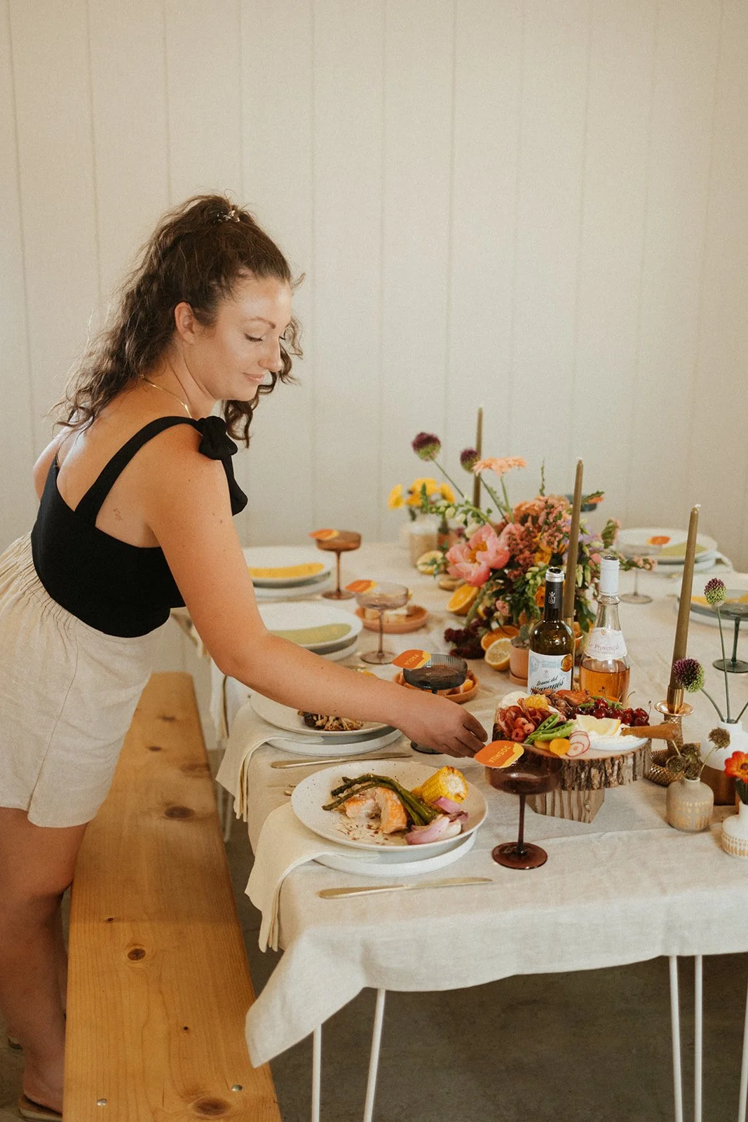 Woman decorating a table with food and flowers, with plates of seafood, drinks, and floral arrangements.
