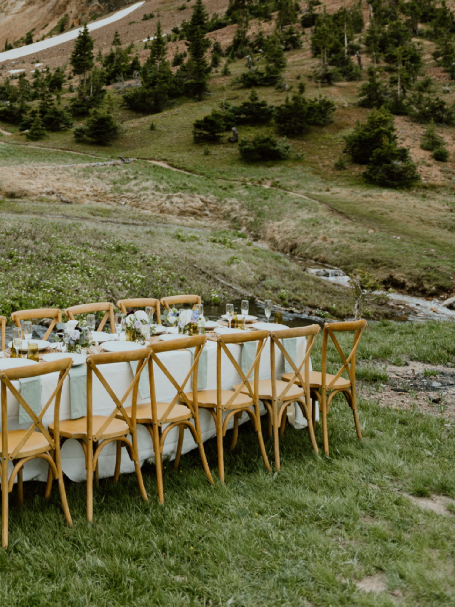 A long dining table set for a meal outdoors on a grassy area with mountain scenery in the background. The table has white tablecloths, floral centerpieces, and glassware, with wooden cross-back chairs arranged along both sides.