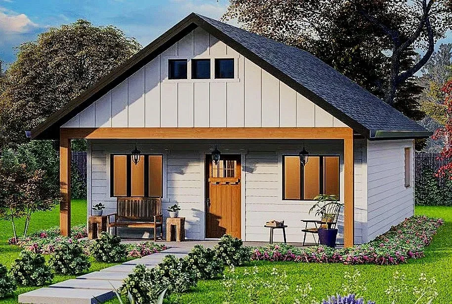A charming white cottage with a dark pitched roof, wooden accents, and a small front porch with outdoor seating, surrounded by lush green grass and colorful flowers.