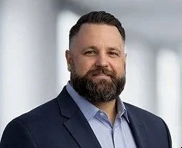 Portrait of a middle-aged man with a beard, wearing a dark suit and light blue shirt, standing against a blurred office background.