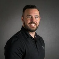 Portrait of a smiling man with short dark hair and a beard, wearing a black polo shirt, against a gray background.